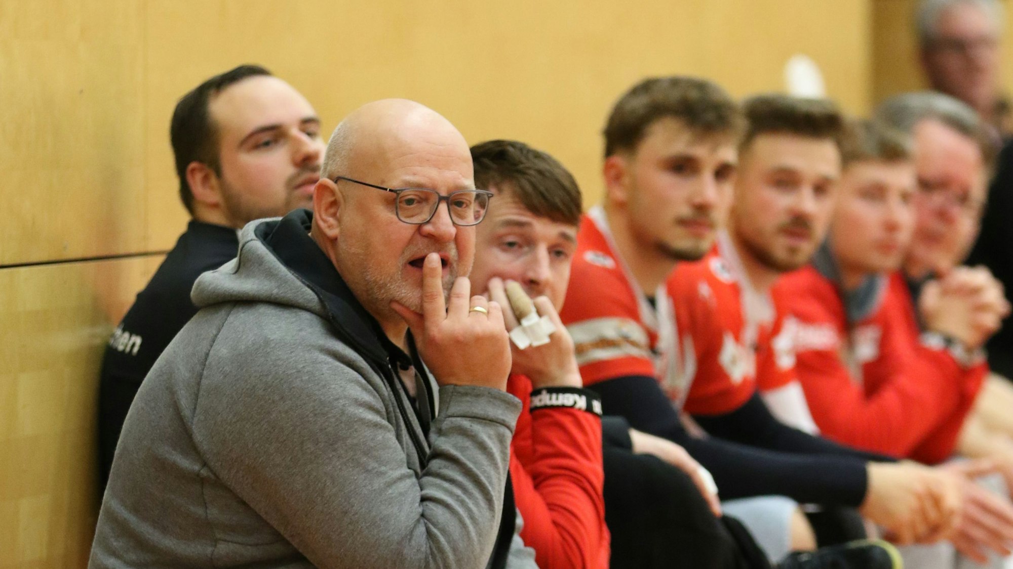 Der Trainer sitzt auf der Spielerbank, im Hintergrund mehrere seiner Akteure.