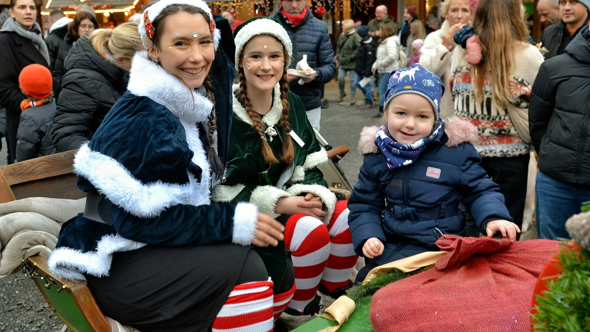 Ein Mädchen mit Anorka und Mütze sitzt auf der Kutsche zweier Elfen, die einen großen roten Geschenkesack dabei haben.