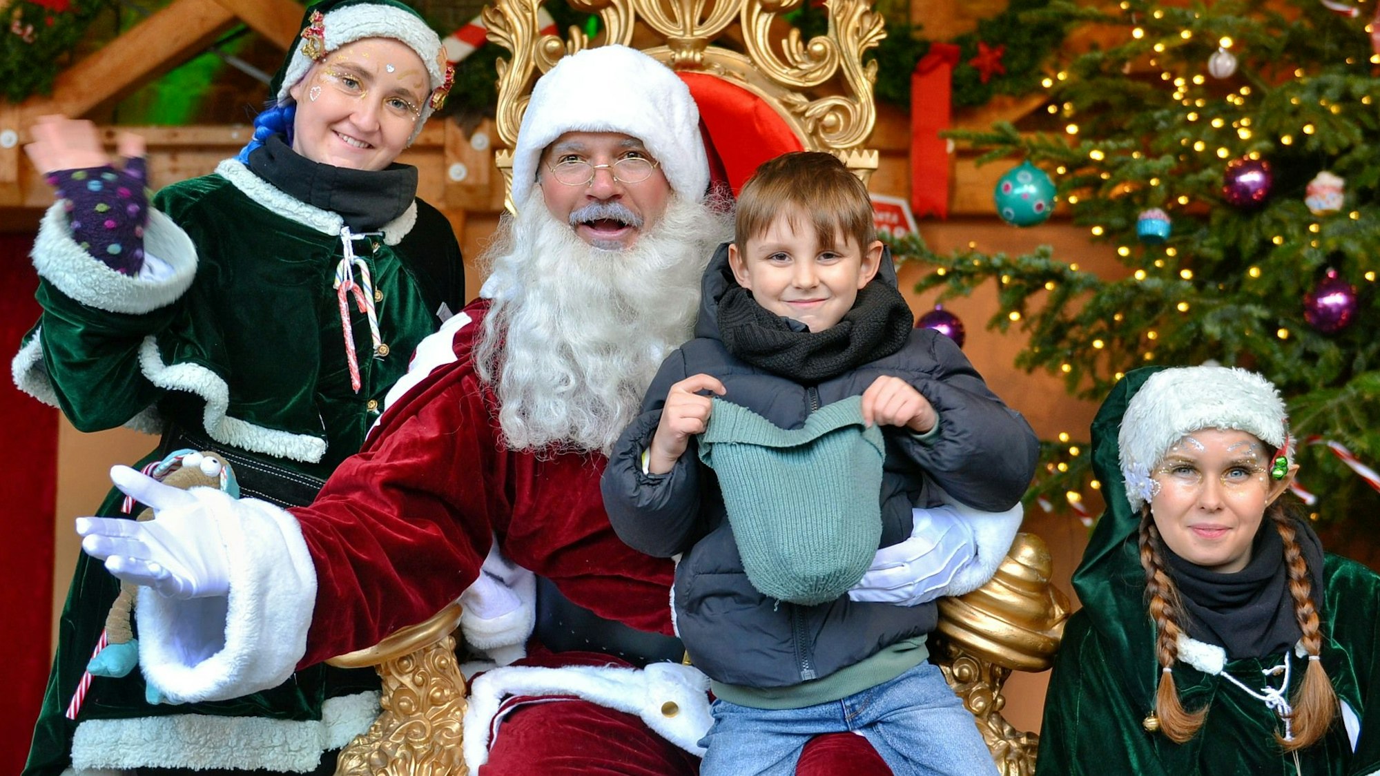 Milian sitzt auf dem Schoß des Weihnachtsmannes, links und rechts daneben haben sich Elfen in blauen Kostümen postiert.