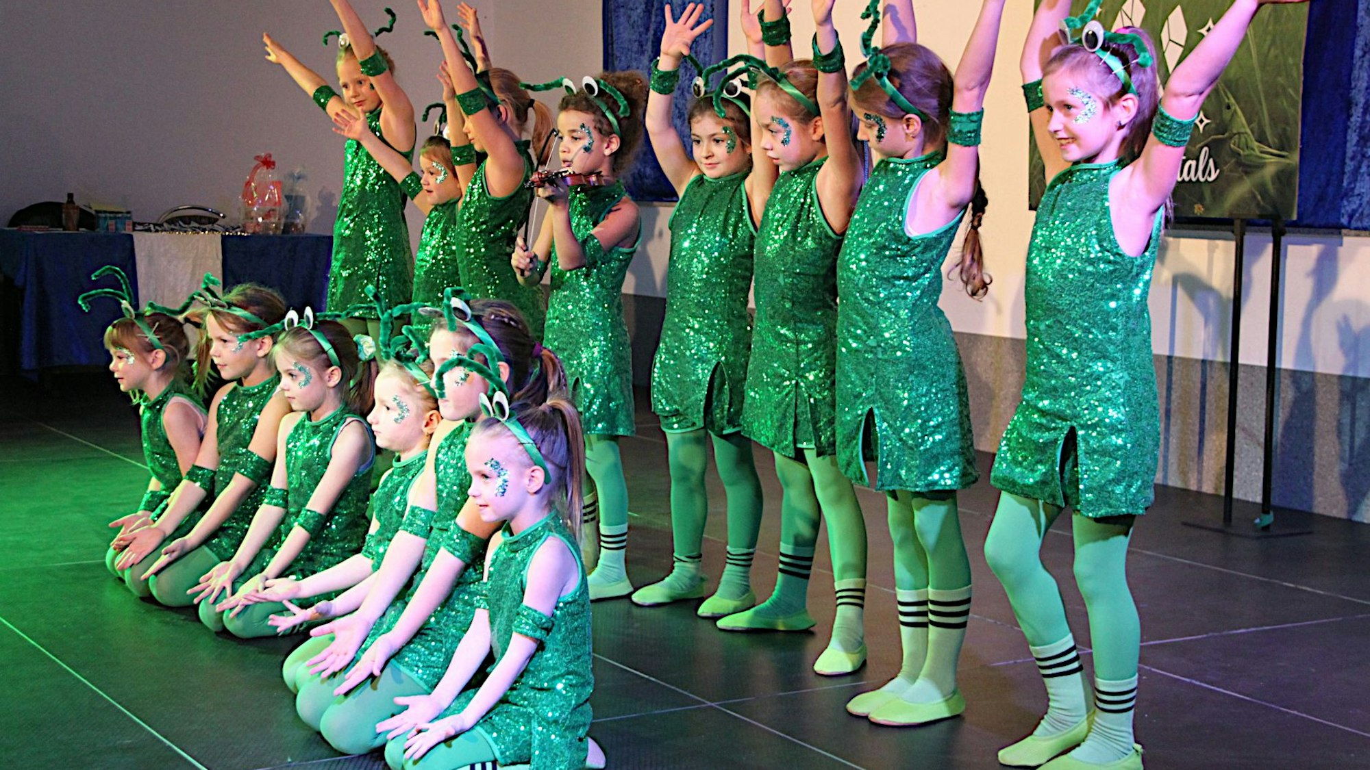 Als grüne Frösche verkleidete Mädchen sitzen und stehen auf der Bühne, einige reißen die Arme nach oben.