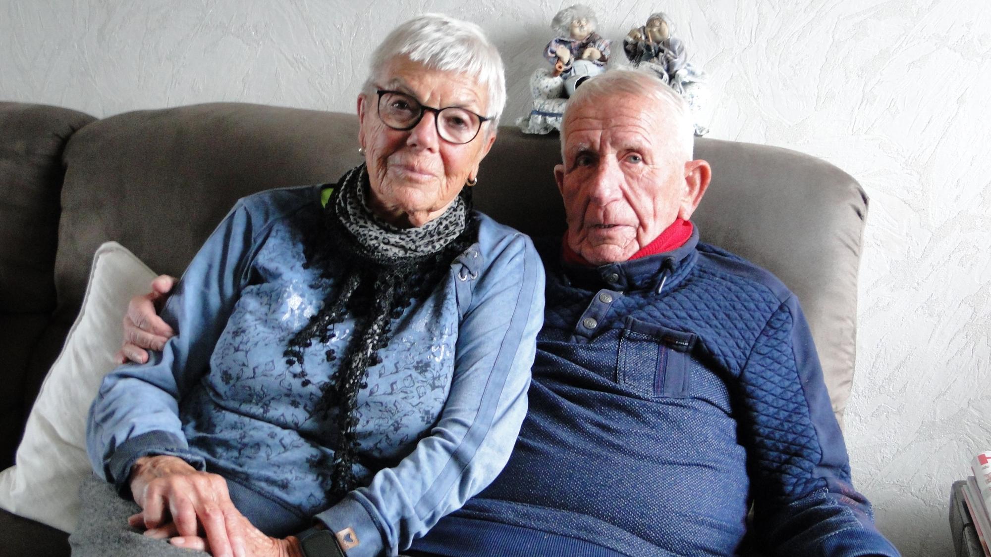 Eine Frau mit kurzem grauen Haar und Brille sitzt neben einem Mann mit grauem Haar auf einem Sofa.