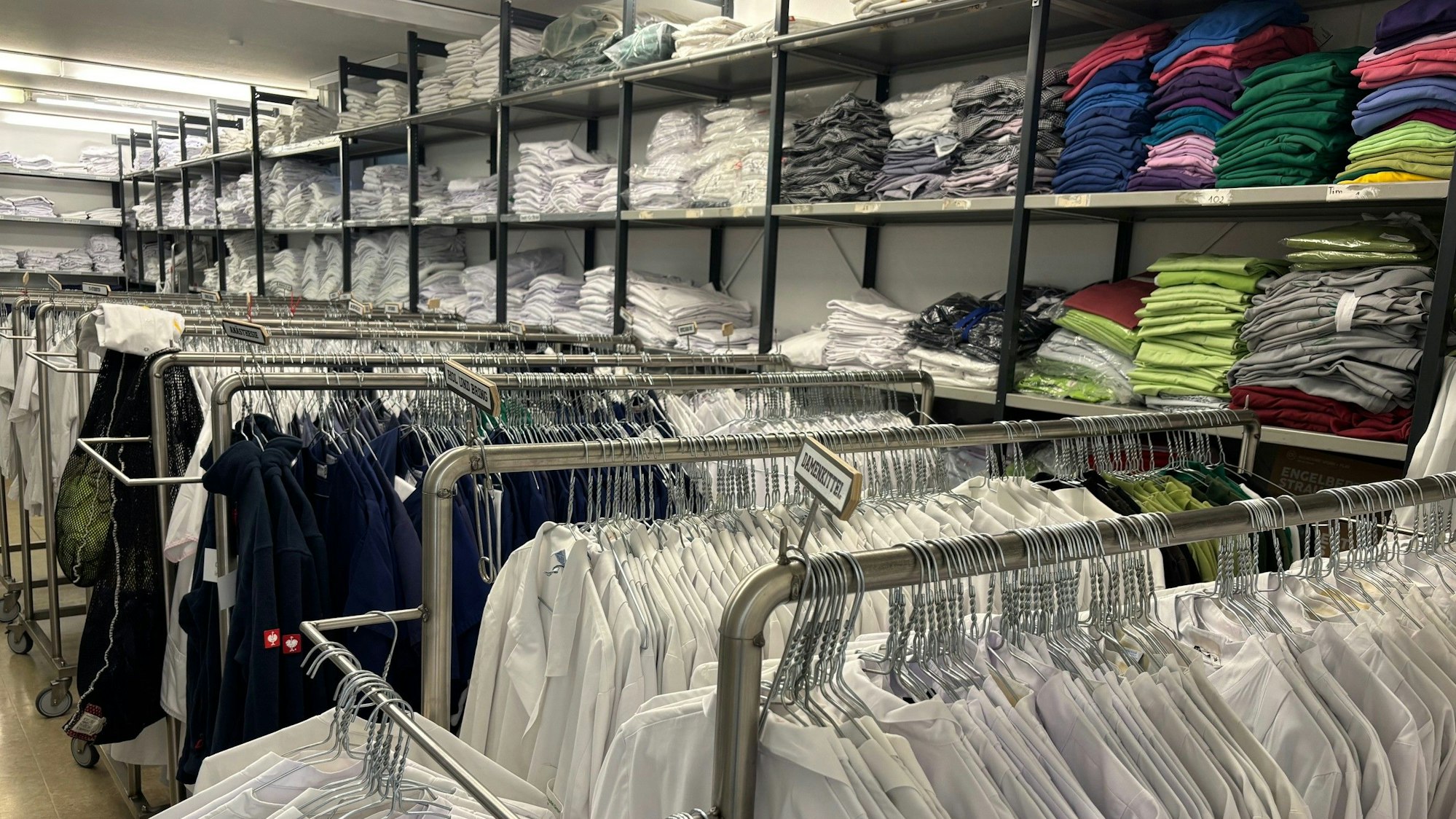 Auf dem Foto sind Stapel von Wäsche zu sehen, auf Kleiderständern hängen Arbeitshemden und -kittel.