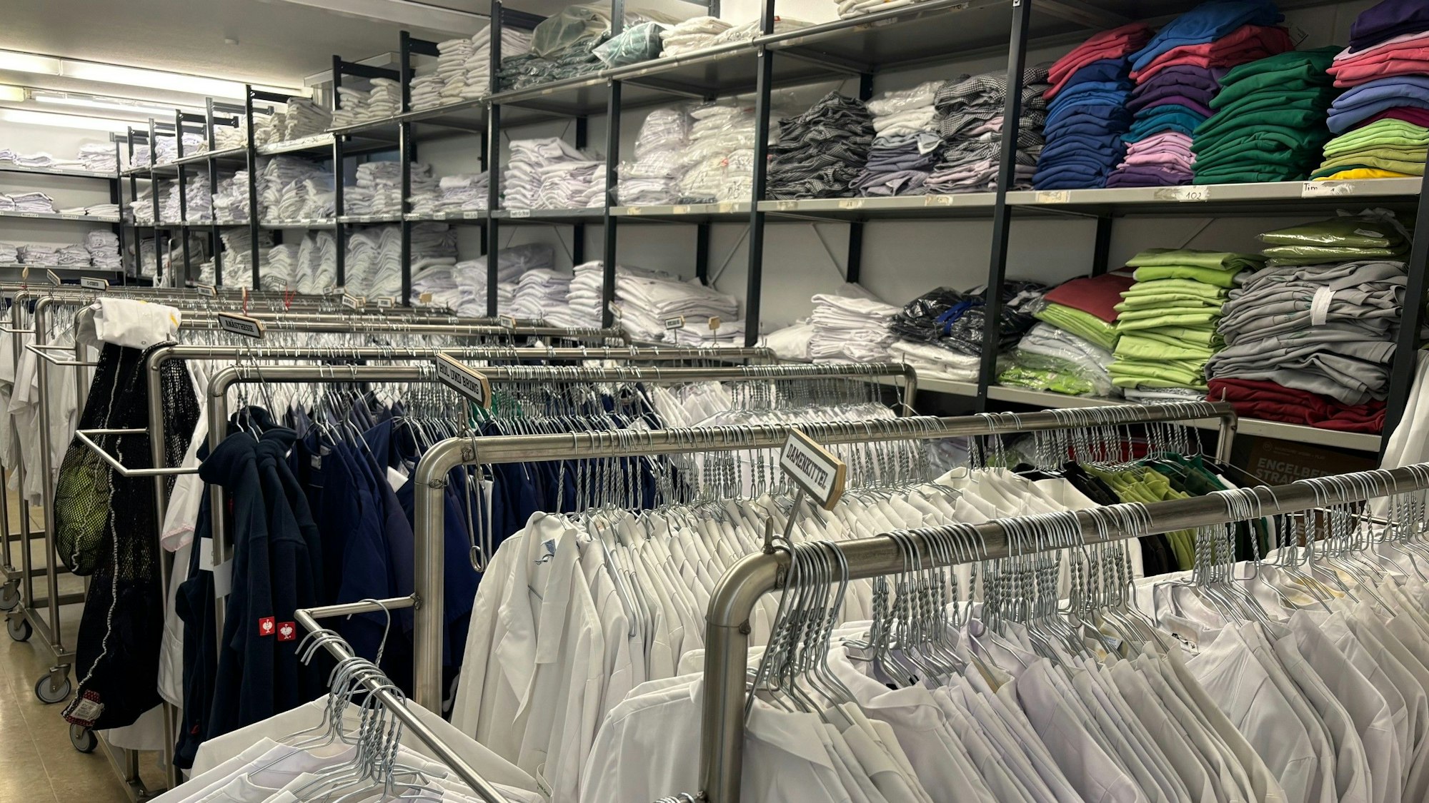 Auf dem Foto sind Stapel von Wäsche zu sehen, auf Kleiderständern hängen Arbeitshemden und -kittel.
