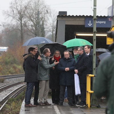 Mehrere Männer und eine Frau auf einem Bahnsteig, im Vordergrund ein Mann mit einer Kamera.