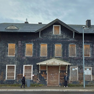 Das Bild zeigt das ehemalige Bahnhofsgebäude. Die Fenster und Türen sind mit Holzplatten zugenagelt.