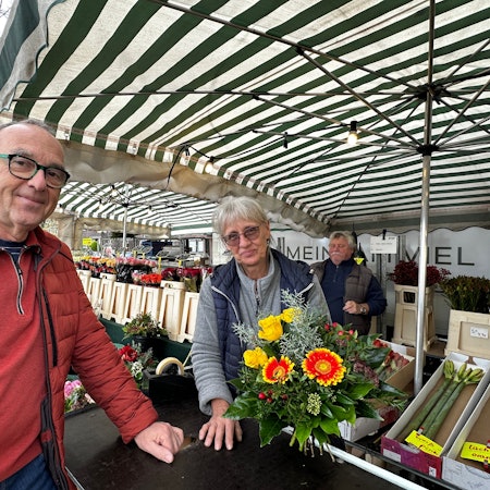 Ein Mann in roter Jacke steht an einem Blumenstand. Die Verkäuferin reicht ihm einen Strauß.