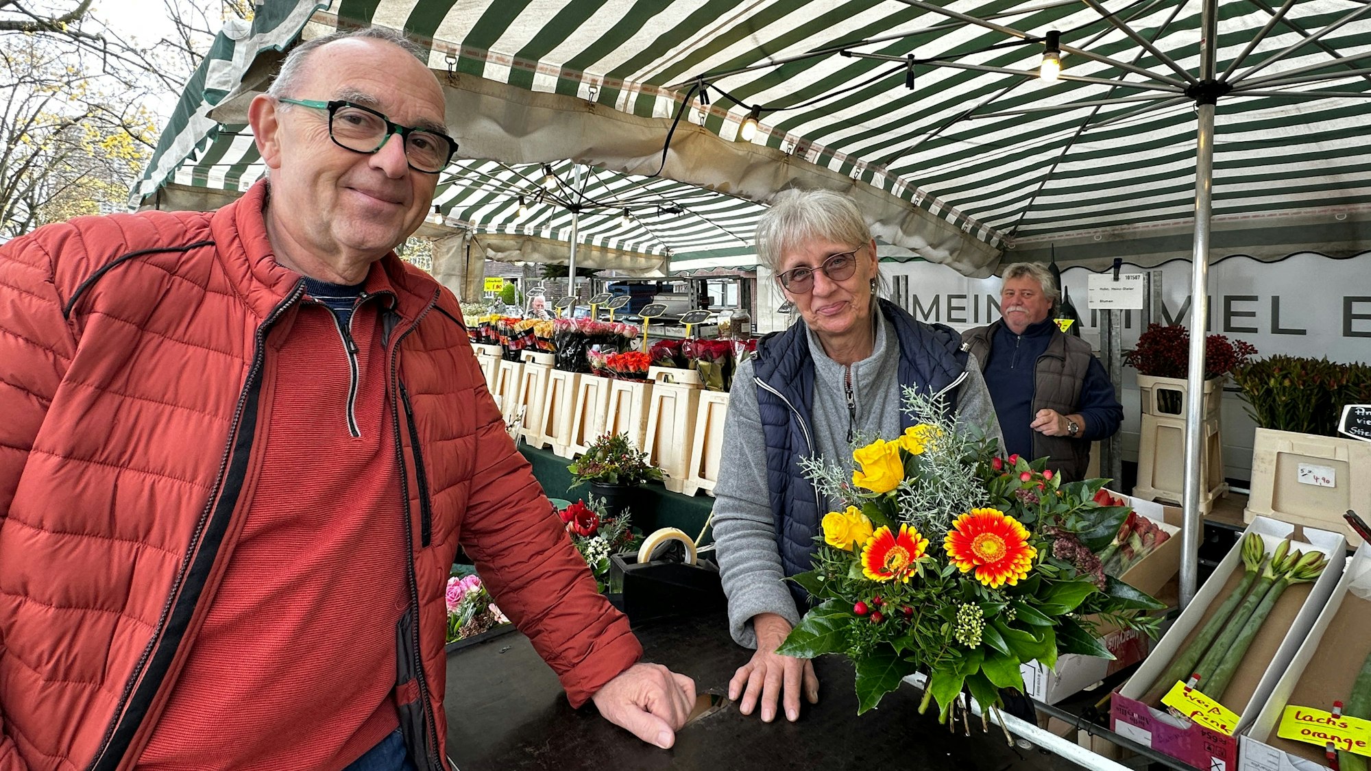Ein Mann in roter Jacke steht an einem Blumenstand. Die Verkäuferin reicht ihm einen Strauß.