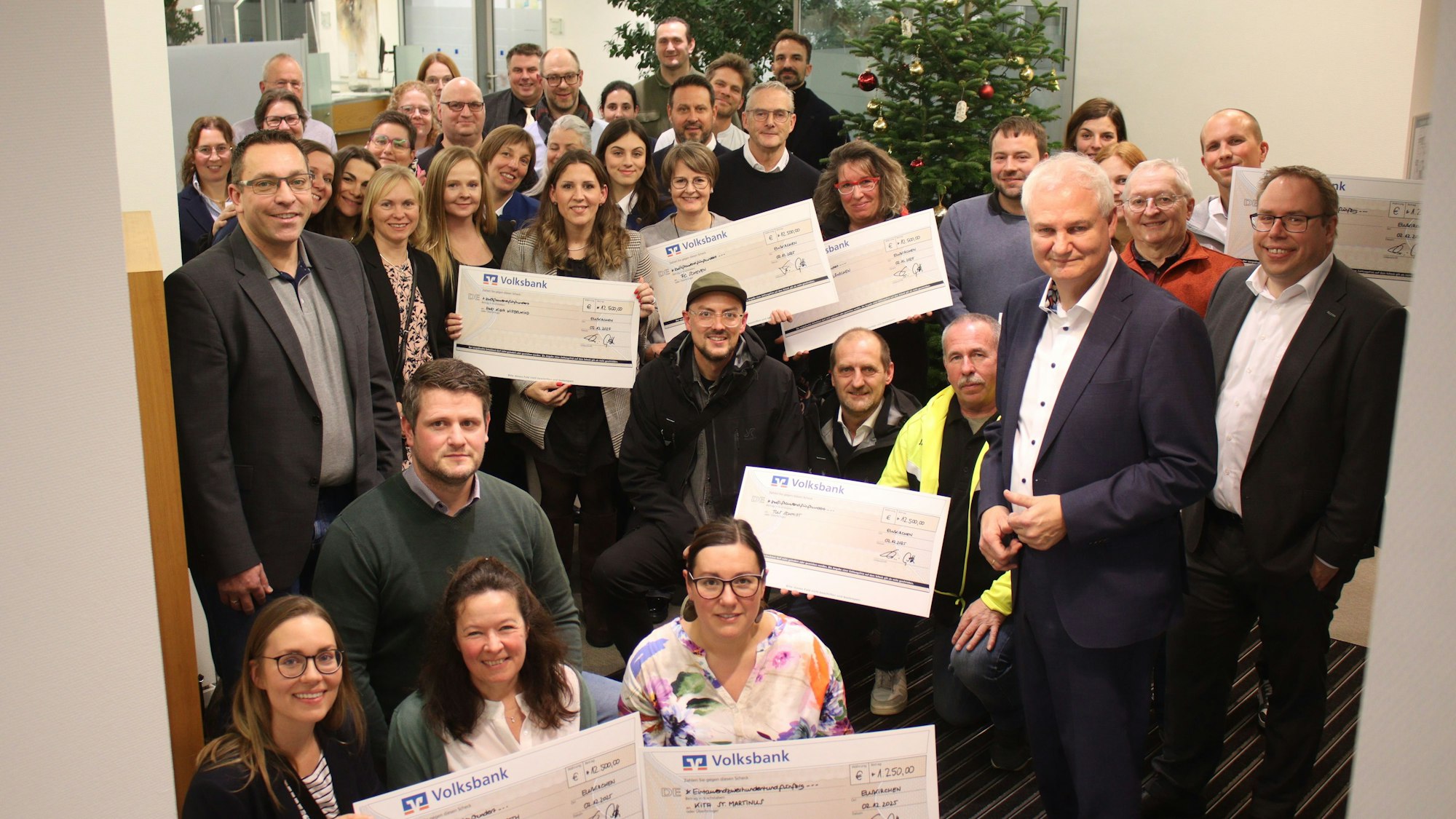 Der Bankenvorstand sowie Vertreter der Organisationen haben sich vor einem Weihnachtsbaum in der Hauptgeschäftsstelle in Euskirchen aufgestellt. Einige halten Schecks in die Höhe.