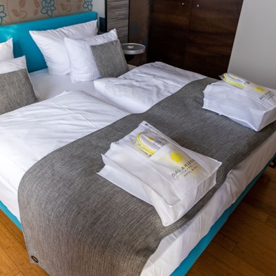 Das Bild zeigt ein Doppelbett in einem Hotel, auf dem zwei Taschen liegen.