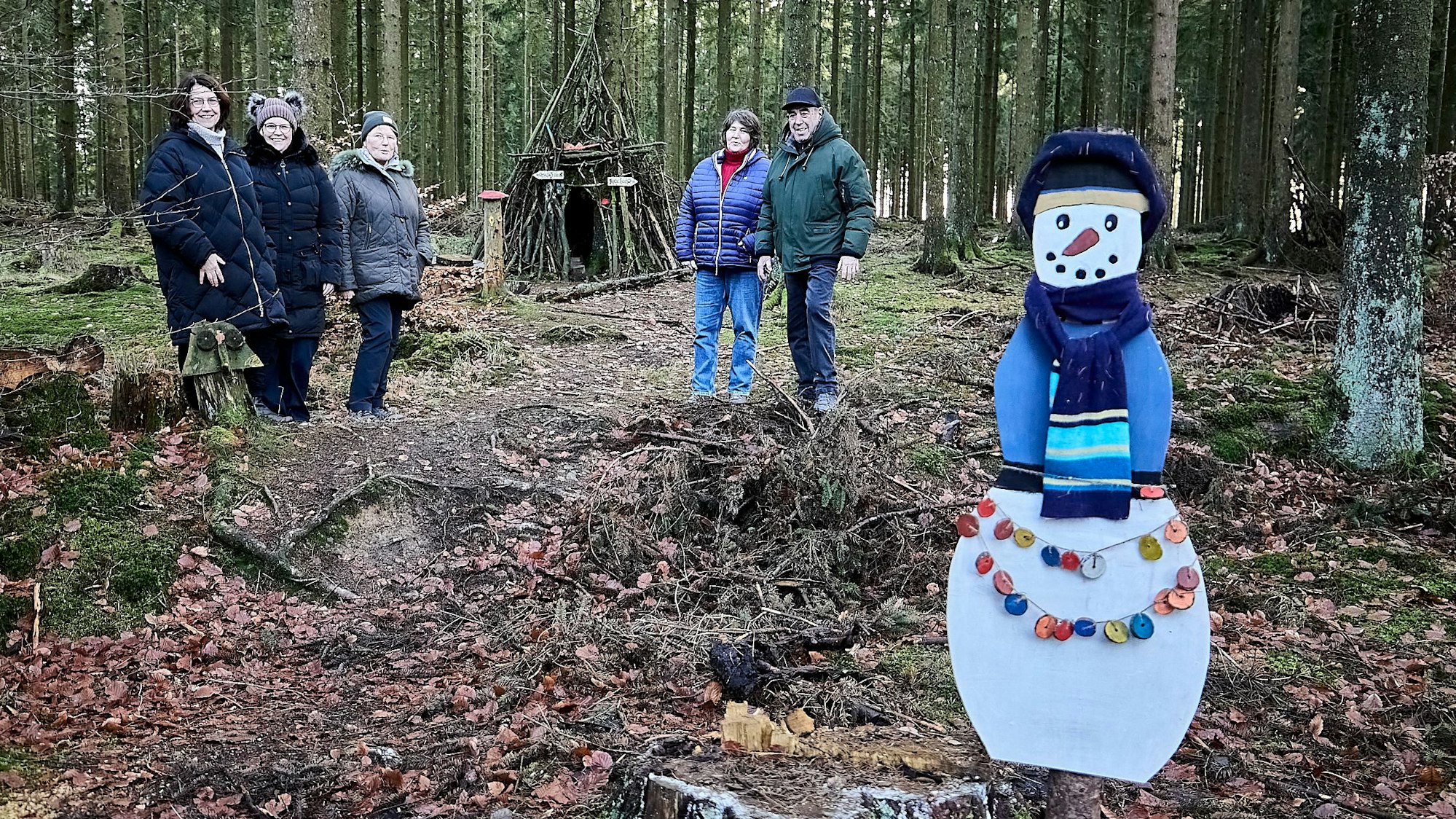 Mitten im Wald stehen die Personen, die den Krippenweg vorgestellt haben. Im Hintergrund sieht man die Schutzhütte, im Vordergrund einen selbstgebastelten Papp-Schneemann mit Schal.