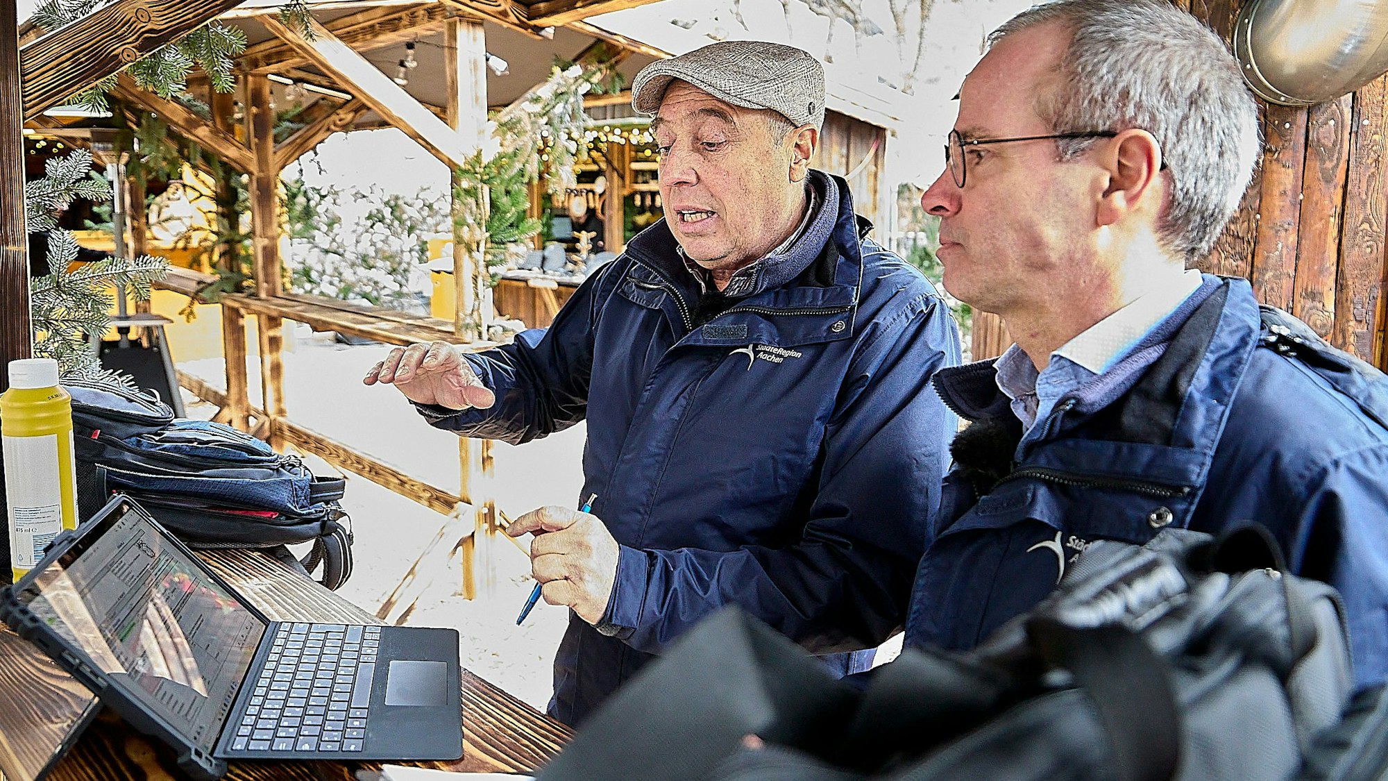 Zwei Männer in blauen Jacken haben einen Laptop auf einen Tresen gestellt und betrachten den Bildschirm.