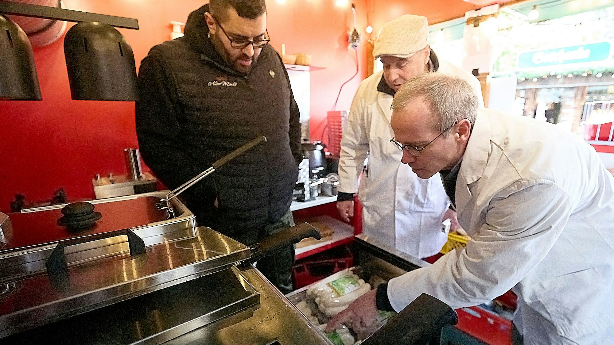 Zwei Lebensmittelkontrolleure in weißen Kitteln stehen in einer Bude des Weihnachtsmarkts in Monschau und überprüfen die in einer Schublade gelagerten Bratwürste.