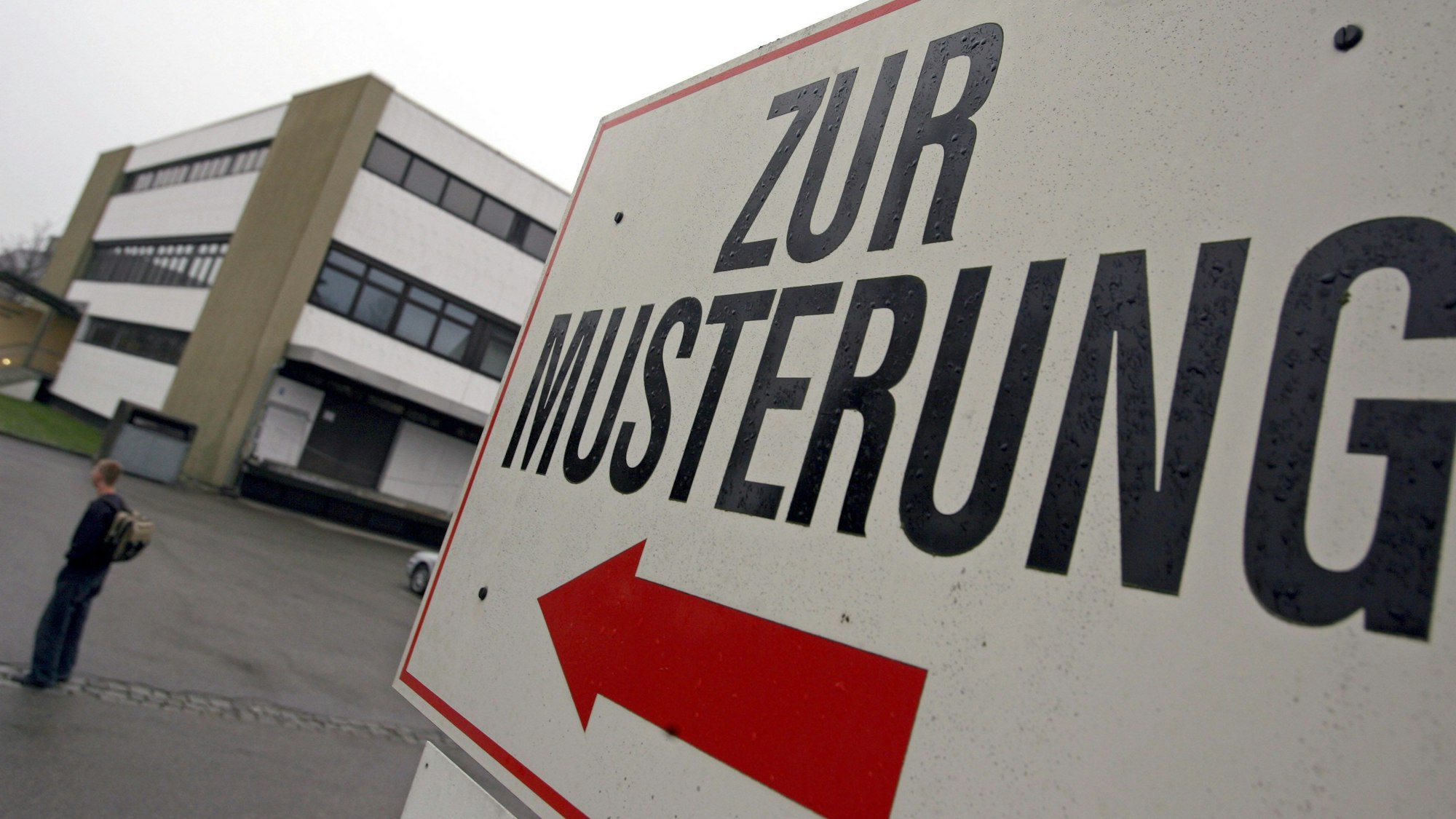 Ein Wehrpflichtiger steht unter einem Hinweisschild mit der Aufschrift «Zur Musterung» vor einem Kreiswehrersatzamt.