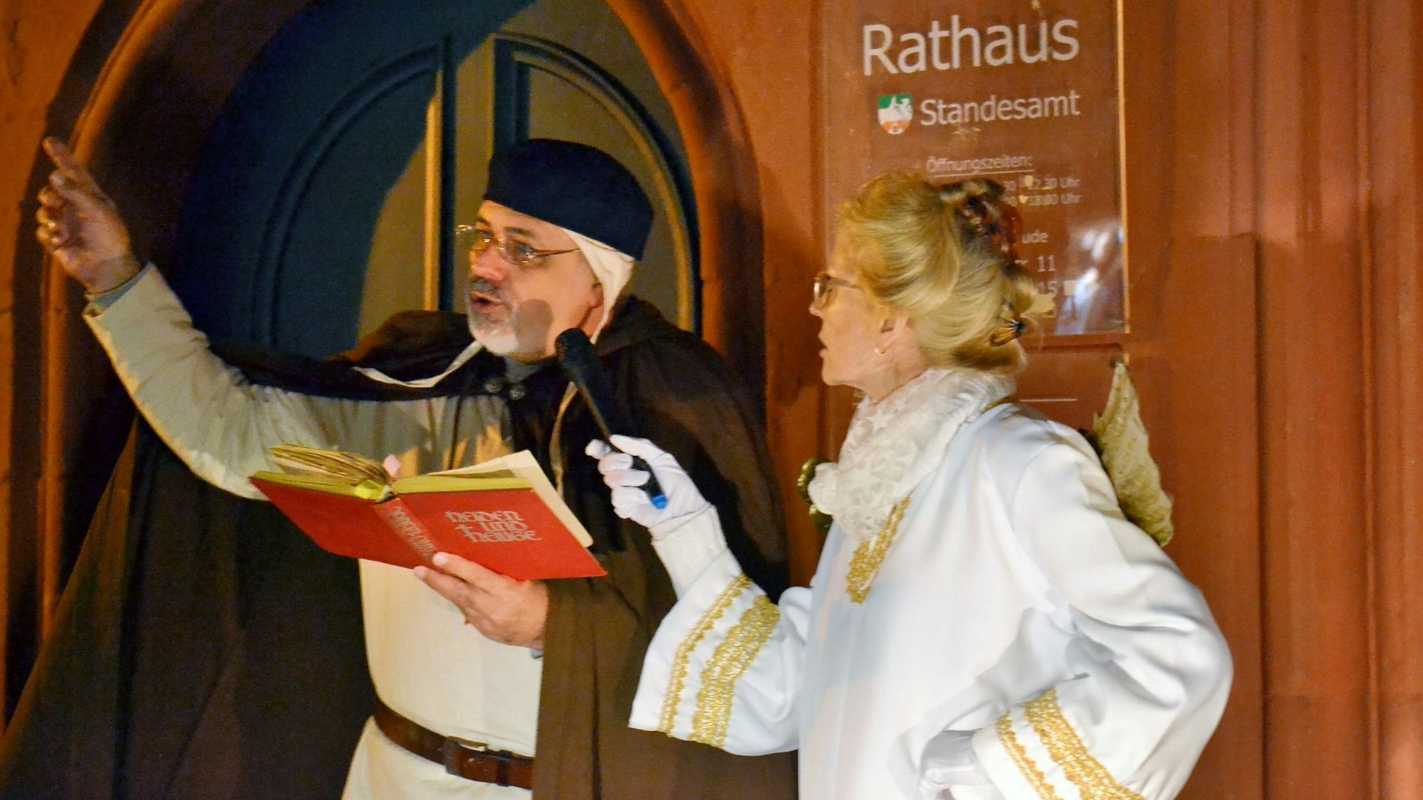 Eine Frau im Engelskostüm hält das Mikrofon für einen Mann, der aus einem roten Buch vorliest.