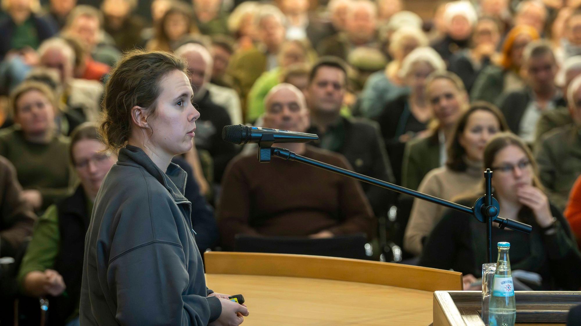 Celin Klimek spricht in ein Mikrofon, hinter ihr sieht man, leicht unsachrf, die dicht gefüllten Zuhörerreihen.