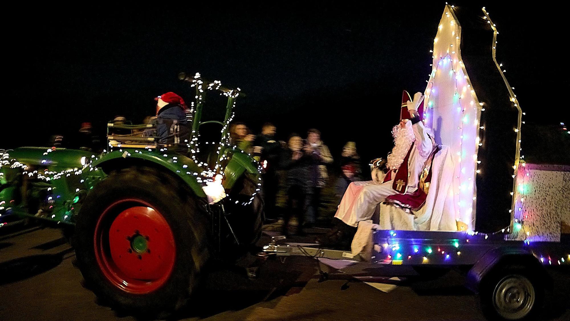 Ein mit Lichterketten geschmückter Trecker zieht einen Anhänger, auf dem der Nikolaus sitzt.