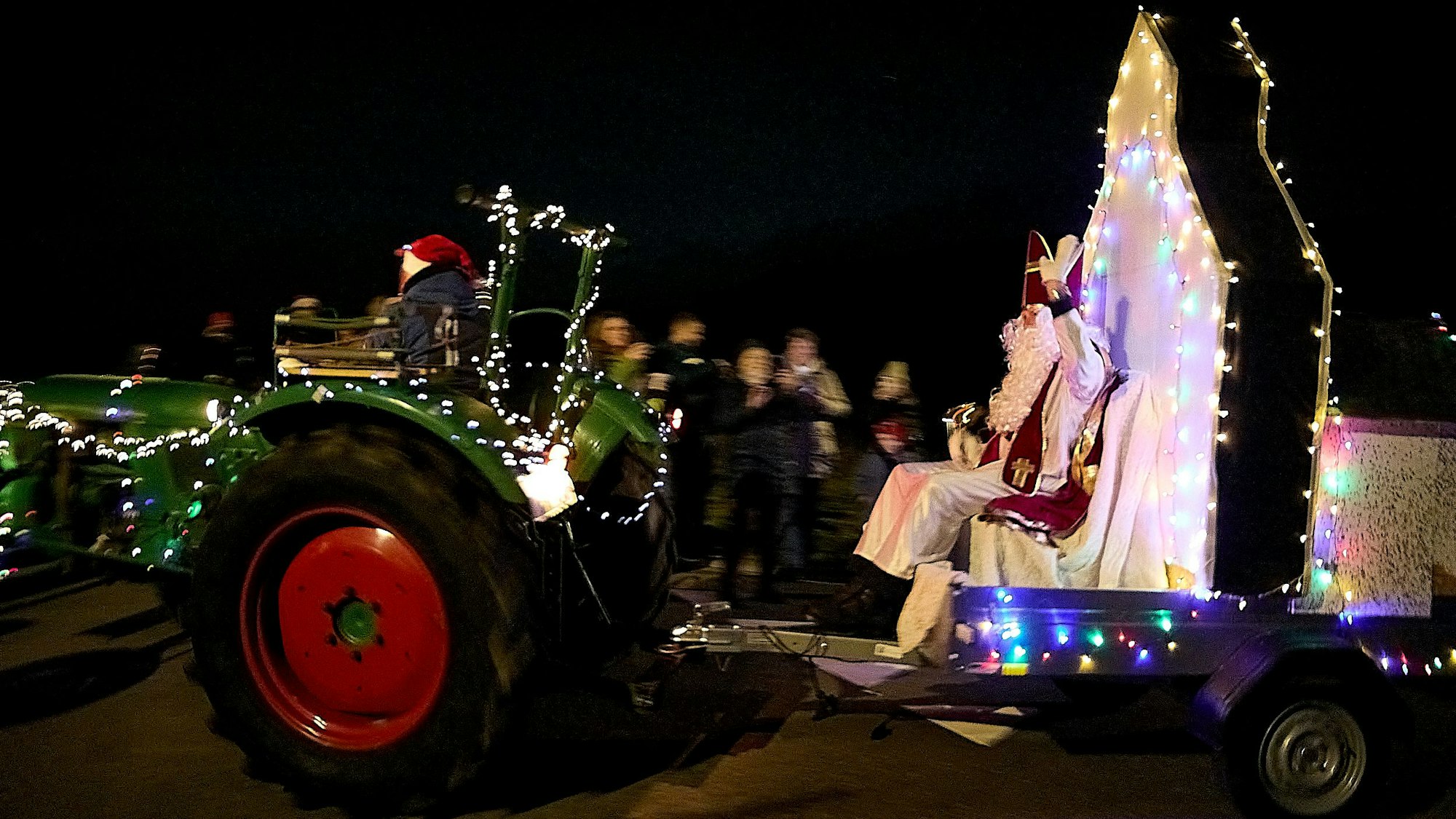 Ein mit Lichterketten geschmückter Trecker zieht einen Anhänger, auf dem der Nikolaus sitzt.
