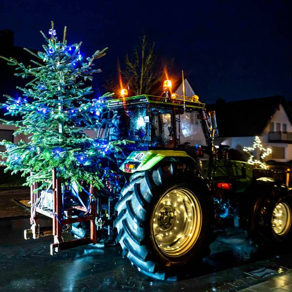 Das Bild zeigt einen Traktor, der einen beleuchteten Weihnachtsbaum transportiert.