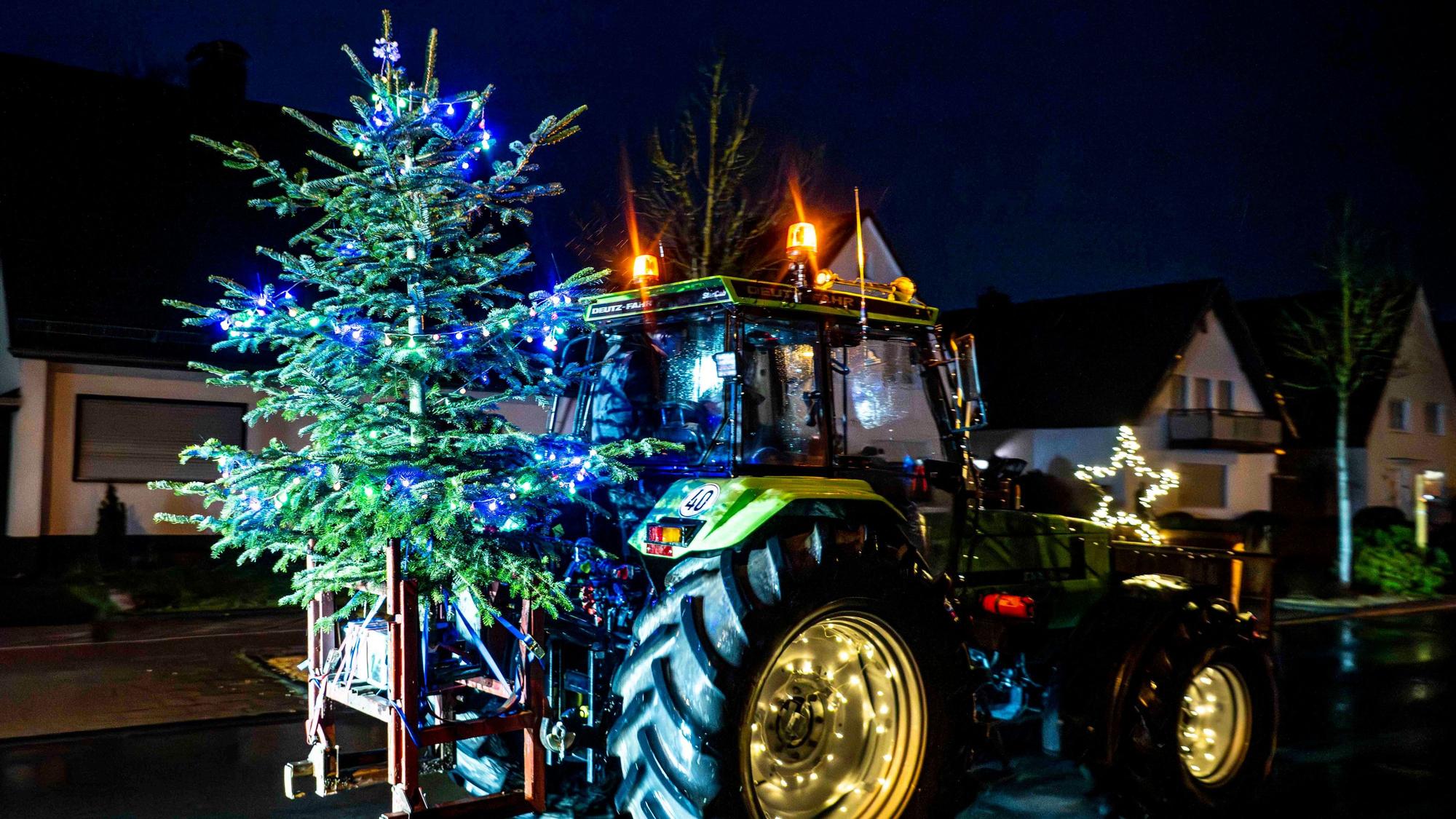 Das Bild zeigt einen Traktor, der einen beleuchteten Weihnachtsbaum transportiert.