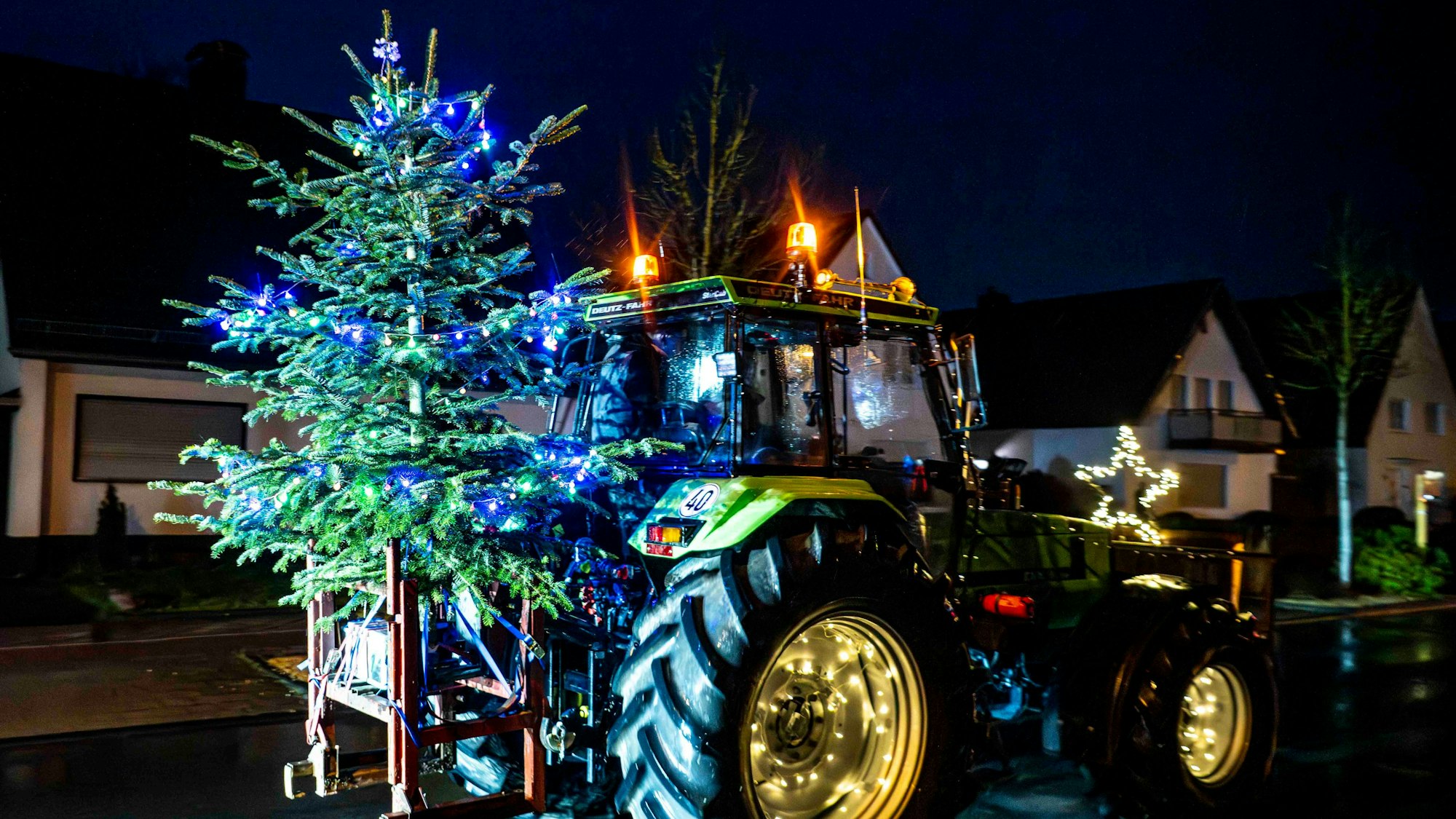 Das Bild zeigt einen Traktor, der einen beleuchteten Weihnachtsbaum transportiert.