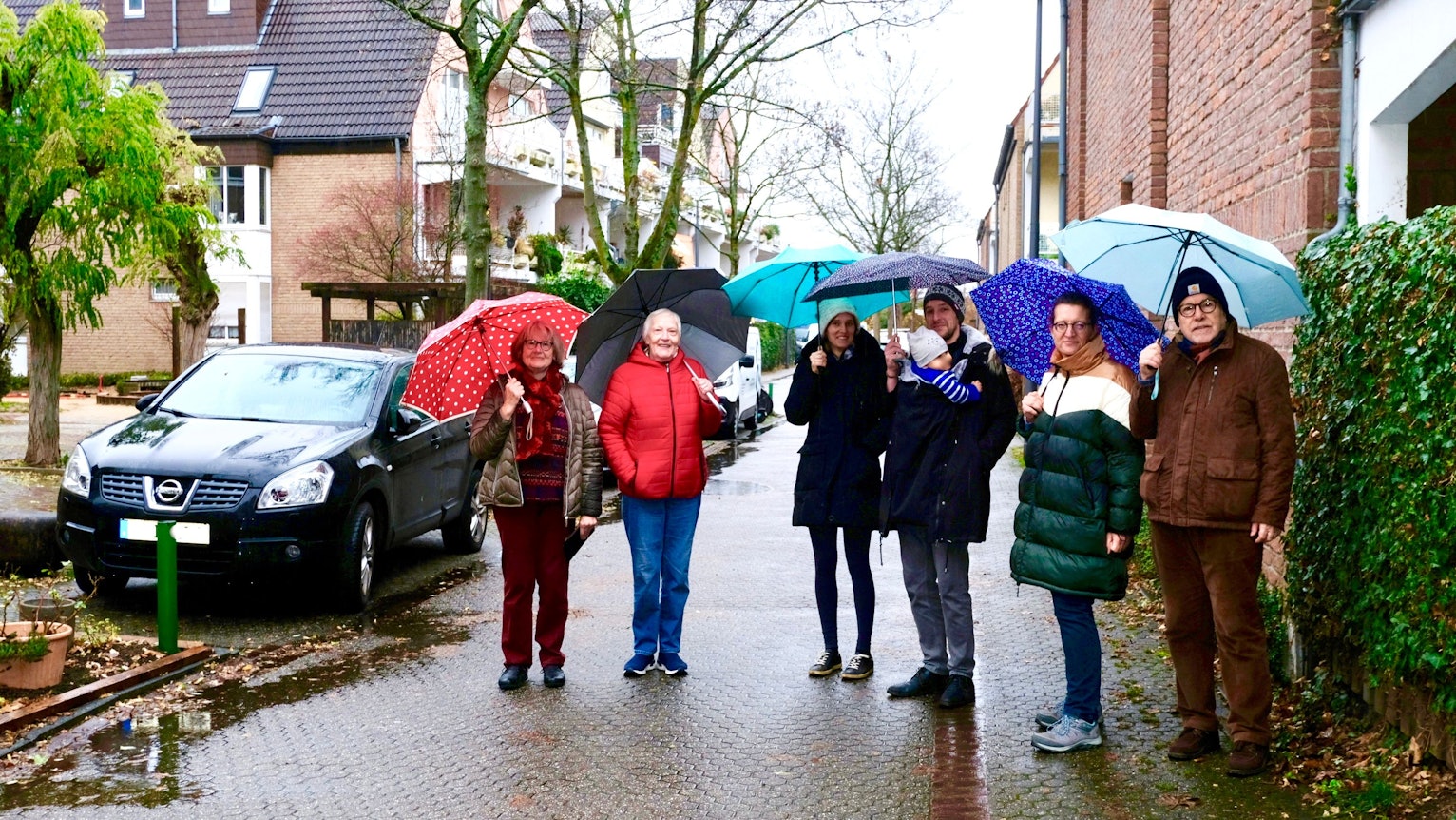 Sechs Personen stehen mit Regenschirmen in einer verkehrsberuhigten Straße. Neben ihnen sind drei Parkplätze besetzt.