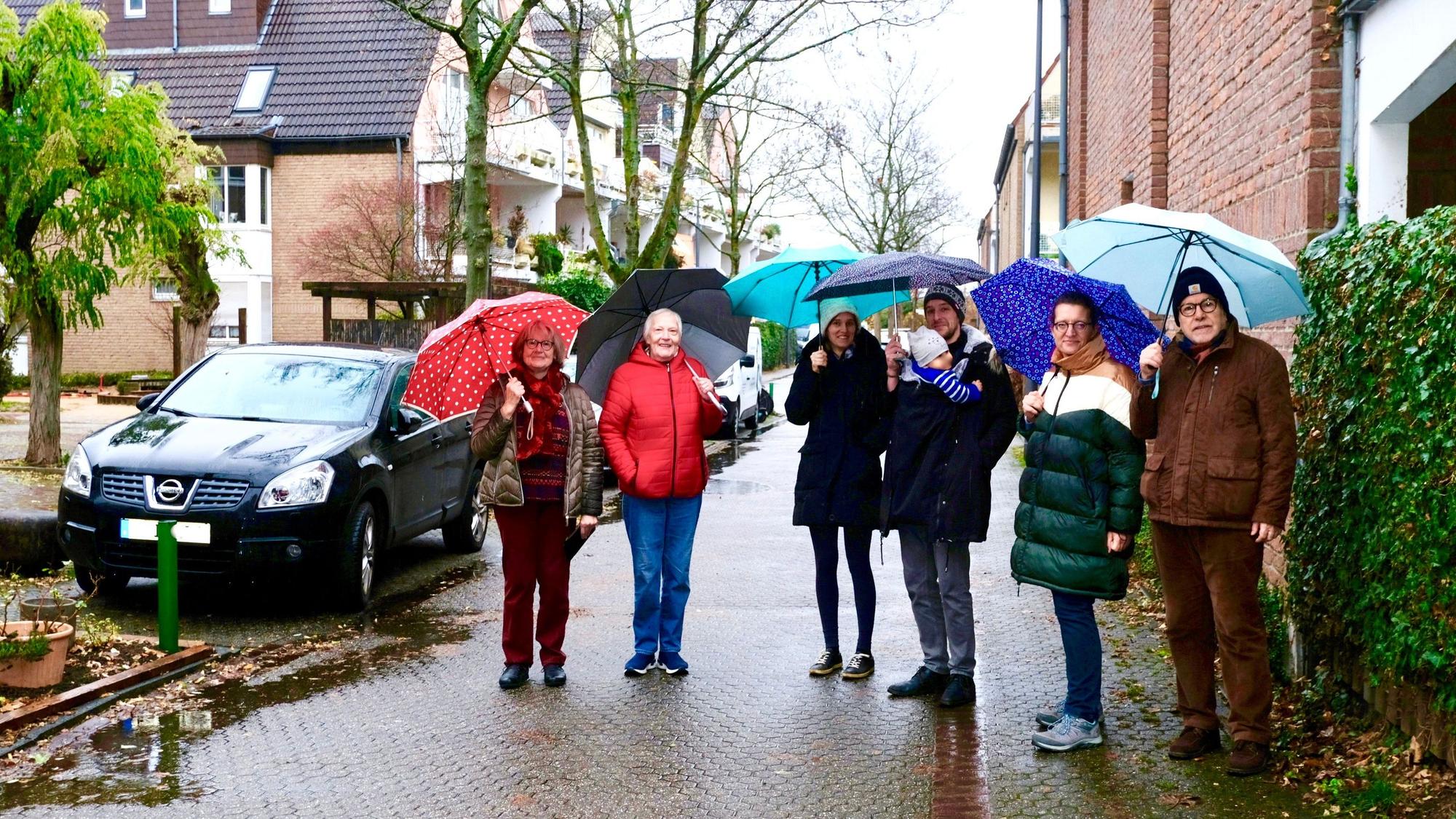 Sechs Personen stehen mit Regenschirmen in einer verkehrsberuhigten Straße. Neben ihnen sind drei Parkplätze besetzt.