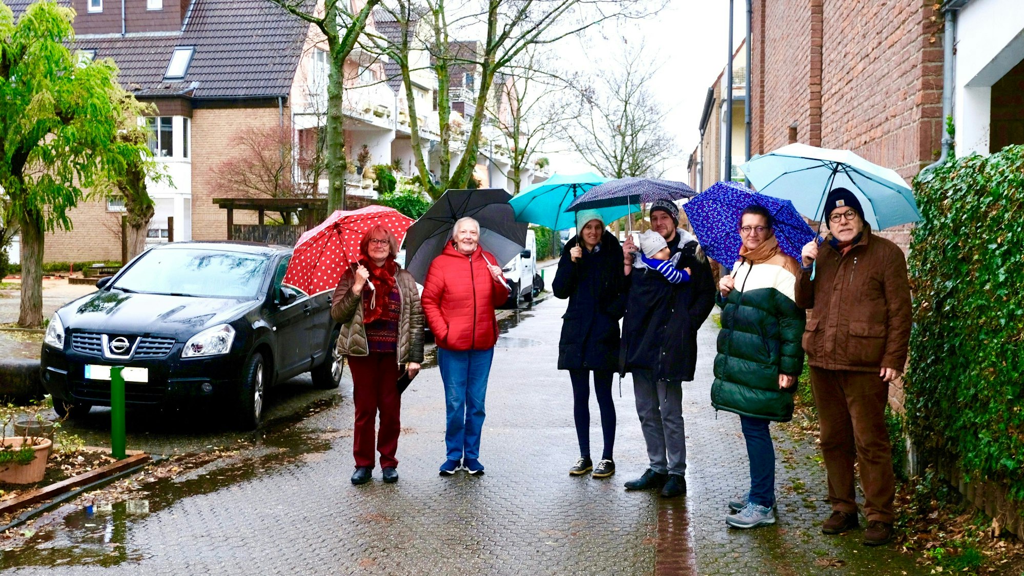 Sechs Personen stehen mit Regenschirmen in einer verkehrsberuhigten Straße. Neben ihnen sind drei Parkplätze besetzt.