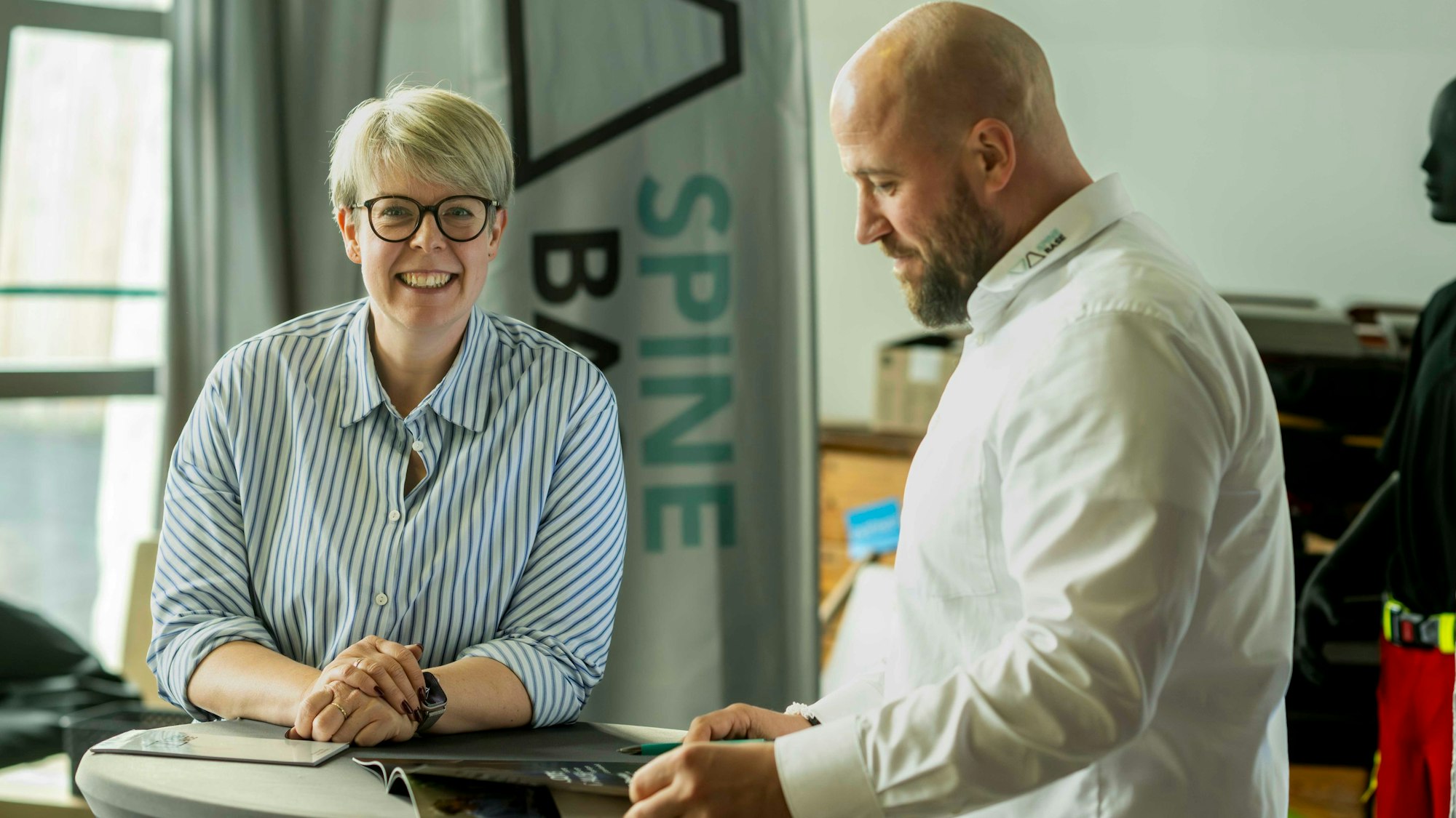 Das Bild zeigt die beiden Obengenannten an einem Stehtisch. Während Neef-Wedler in die Kamera schaut, blättert Gissinger in einem Prospekt.