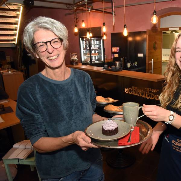 Ulrike Schuster und Bernice Schröder lachen in die Kamera, während sie einen Kaffee und ein Stück Kuchen auf einem Tablett halten.