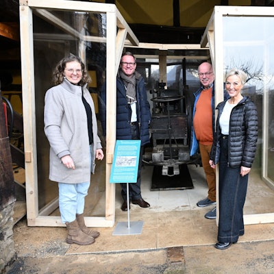 Zwei Frauen und zwei Männer stehen vor einer kleinen historischen Lokomobile, die in einem Glaskasten steht.