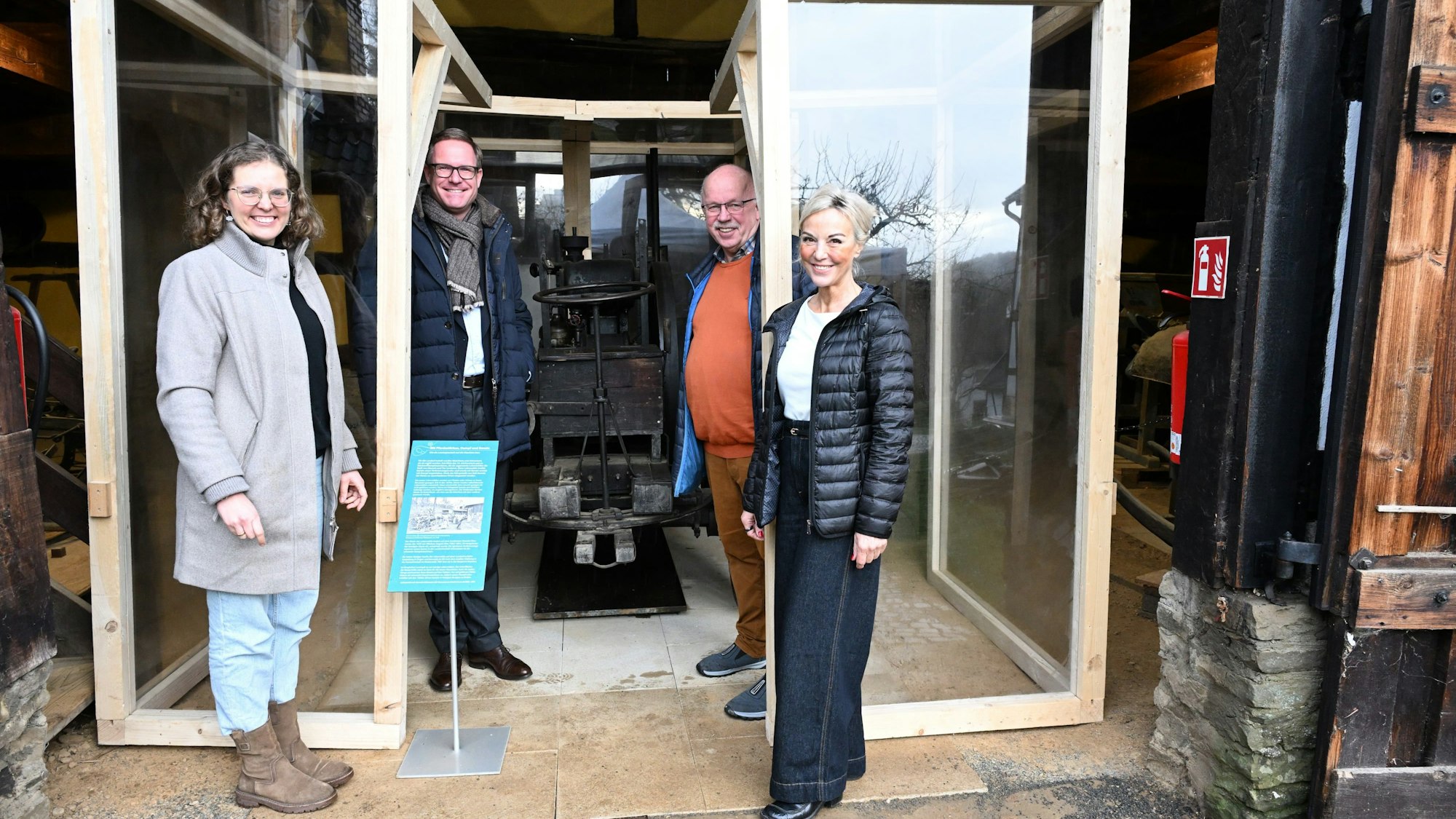 Zwei Frauen und zwei Männer stehen vor einer kleinen historischen Lokomobile, die in einem Glaskasten steht.