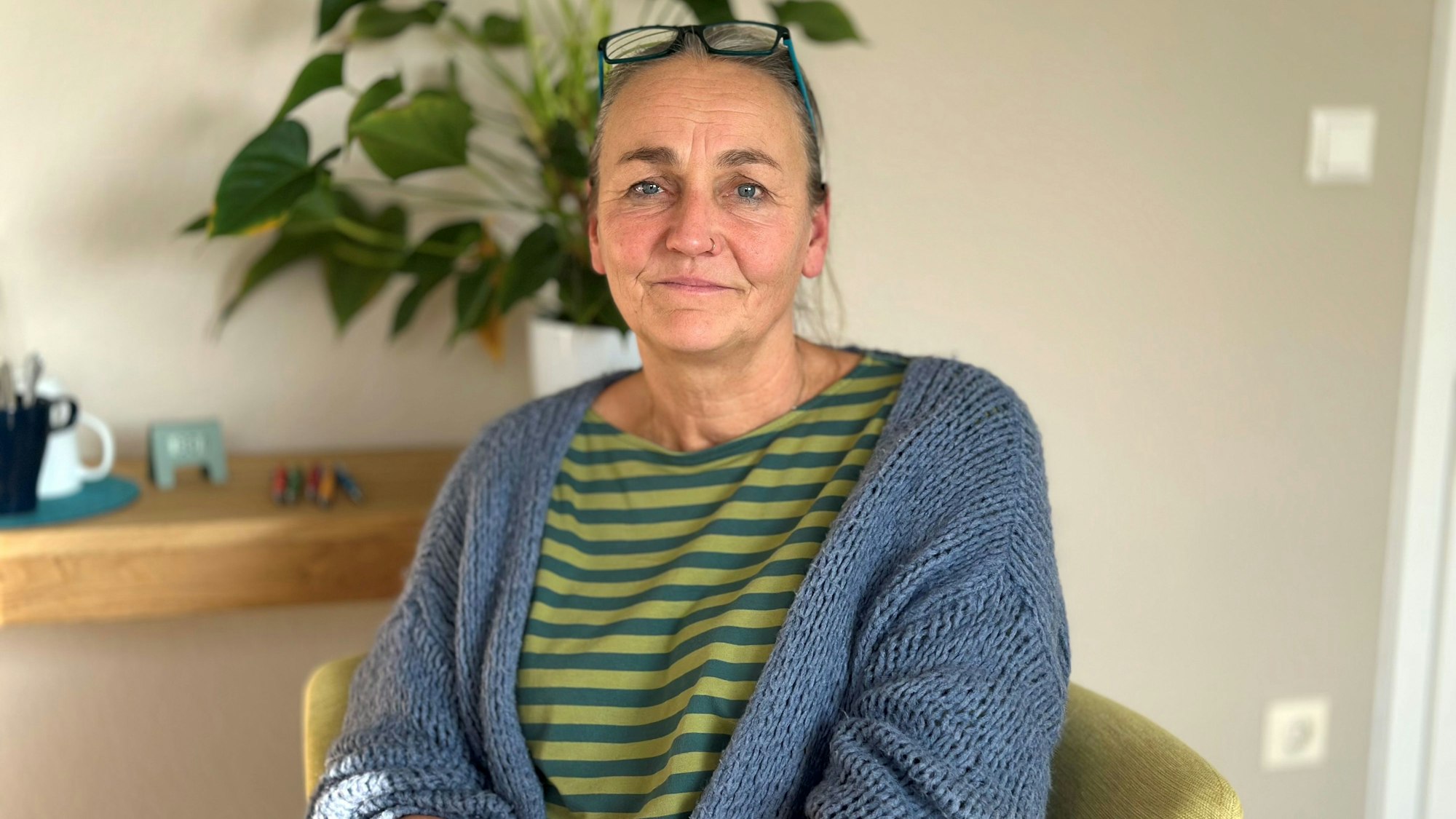 Eine Frau sitzt auf einem gelben Sessel in einem hellen Raum, hinter ihr steht eine Pflanze auf einem Regalbrett.