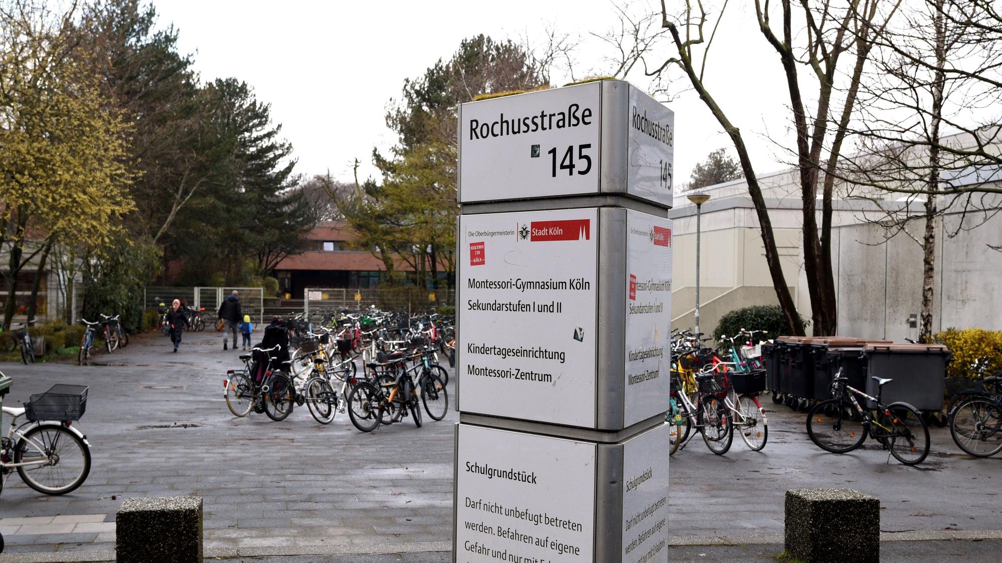 Auf einer Infosäule steht Montessori-Gymnasium, dahinter sind der Vorplatz der Schule, rechts die Turnhalle und im Hintergrund das Schulgebäude zu sehen.