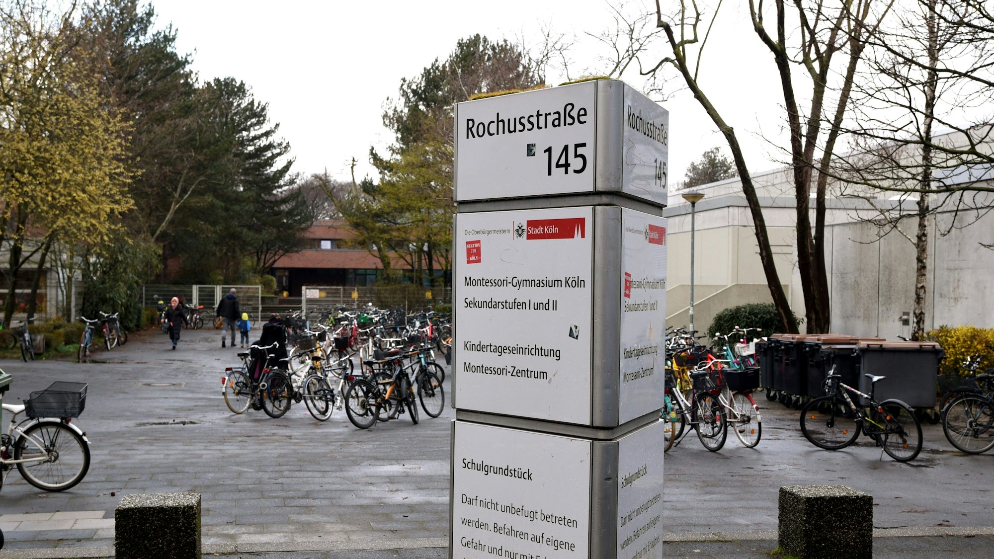 Auf einer Infosäule steht Montessori-Gymnasium, dahinter sind der Vorplatz der Schule, rechts die Turnhalle und im Hintergrund das Schulgebäude zu sehen.