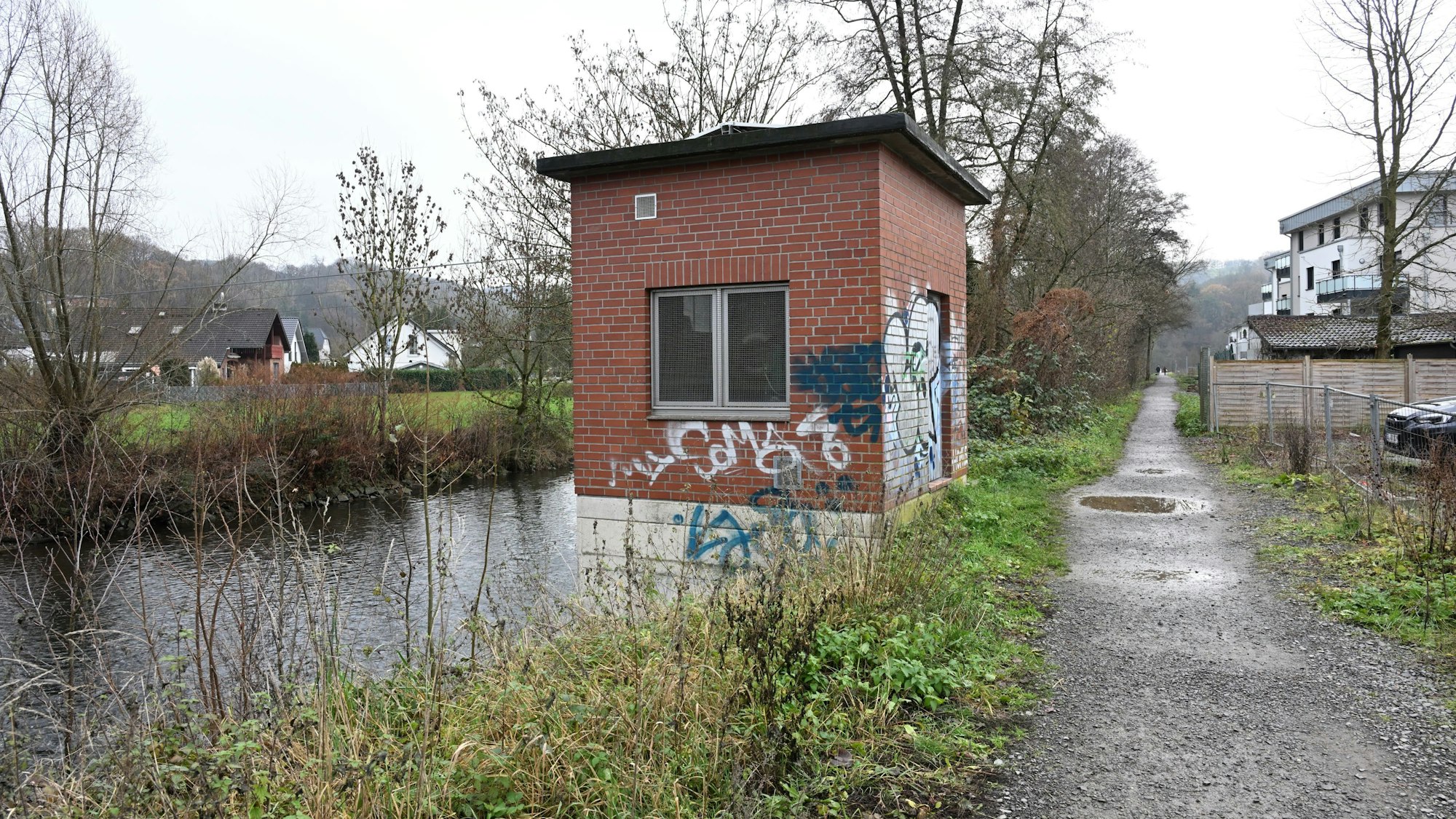 Zu sehen ist ein Fluss, daneben ein Schotterweg und ein Pegelhaus aus roten Backsteinen.