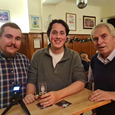 Drei Männer stehen an eine Theke, vor dem Mann ganz links steht ein Aufnahmegerät auf der Theke.