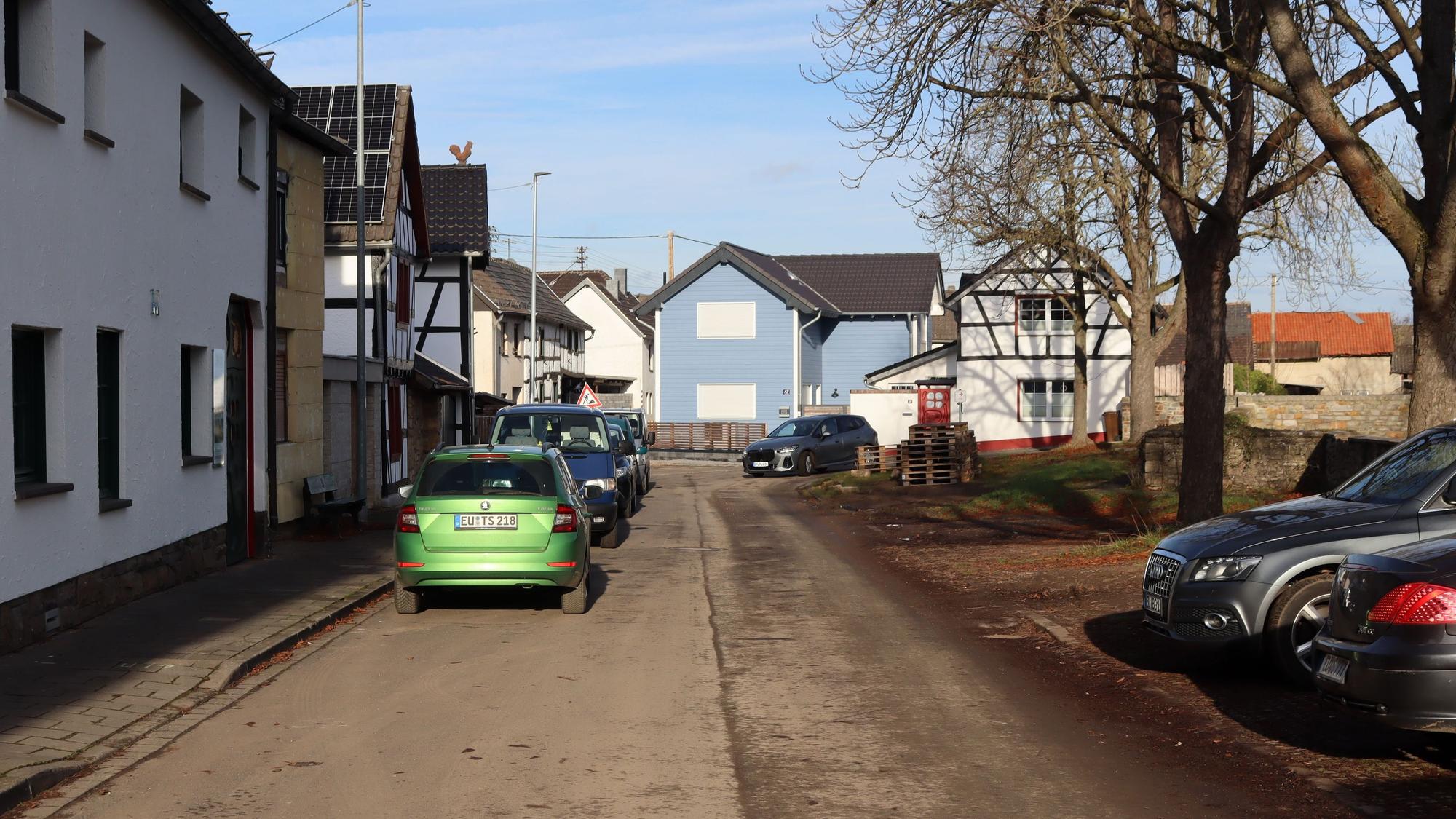 Ein Blick in die Straße Am Bloch in Iversheim. Rechts auf der Grünfläche stehen Autos quer, links vor den Häusern parken Autos längs.