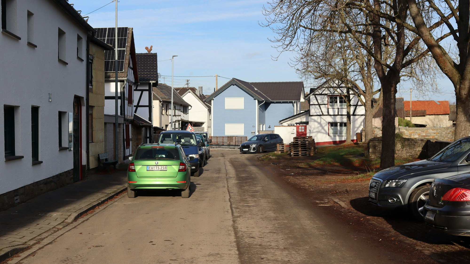 Ein Blick in die Straße Am Bloch in Iversheim. Rechts auf der Grünfläche stehen Autos quer, links vor den Häusern parken Autos längs.