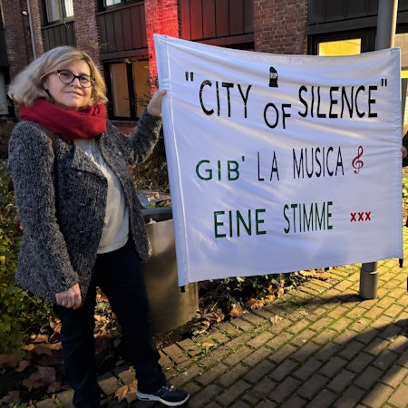 Das Foto zeigt zwei Menschen, die ein Plakat mit der Aufschrift „City of Silence Gib La Musica eine Stimme“ beschriftet ist.