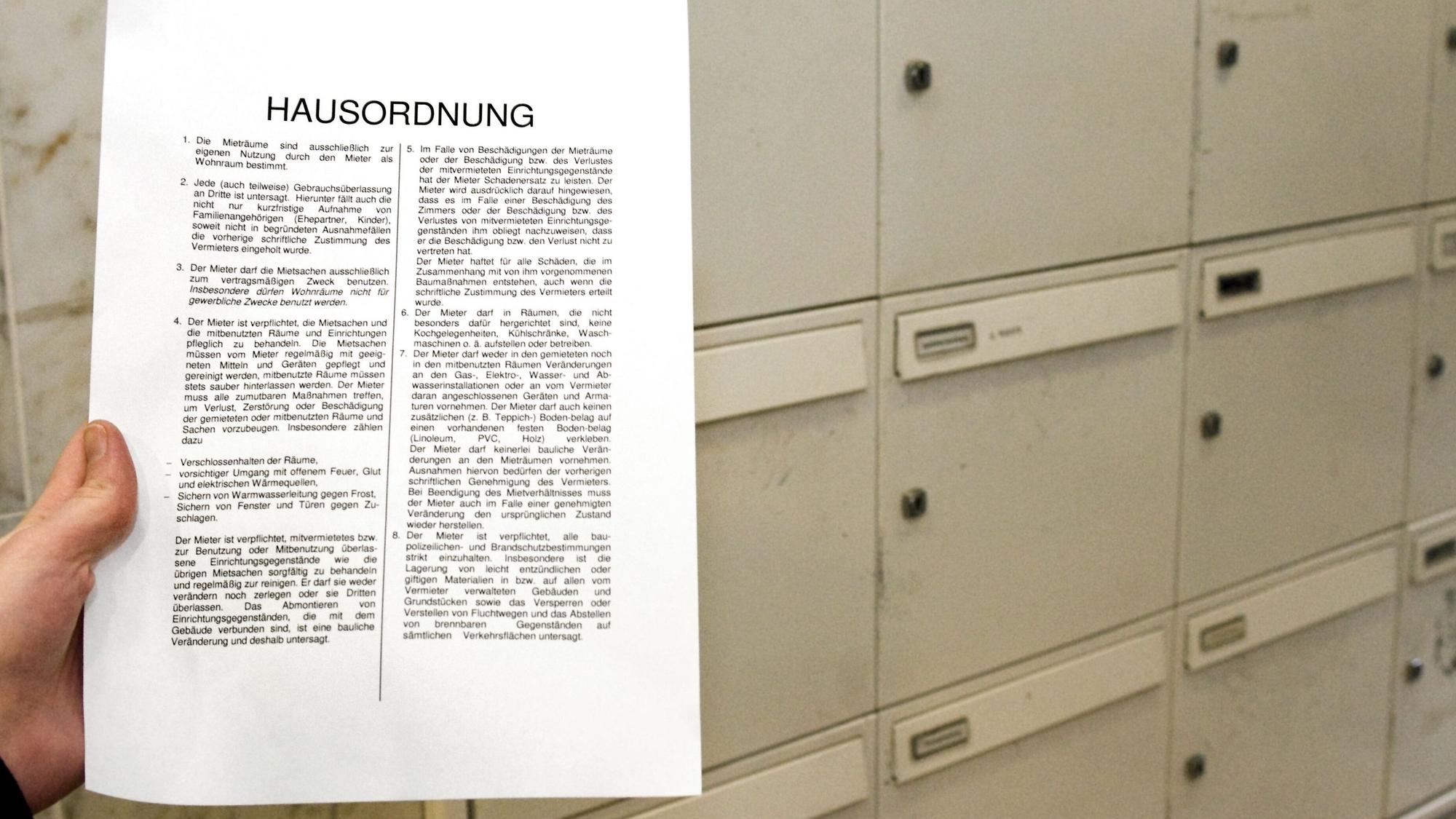 Zu sehen ist einen Hand, die ein Blatt mit der Überschrift Hausordnung im Flur eines Mehrfamilienhauses in die Kamera hält.