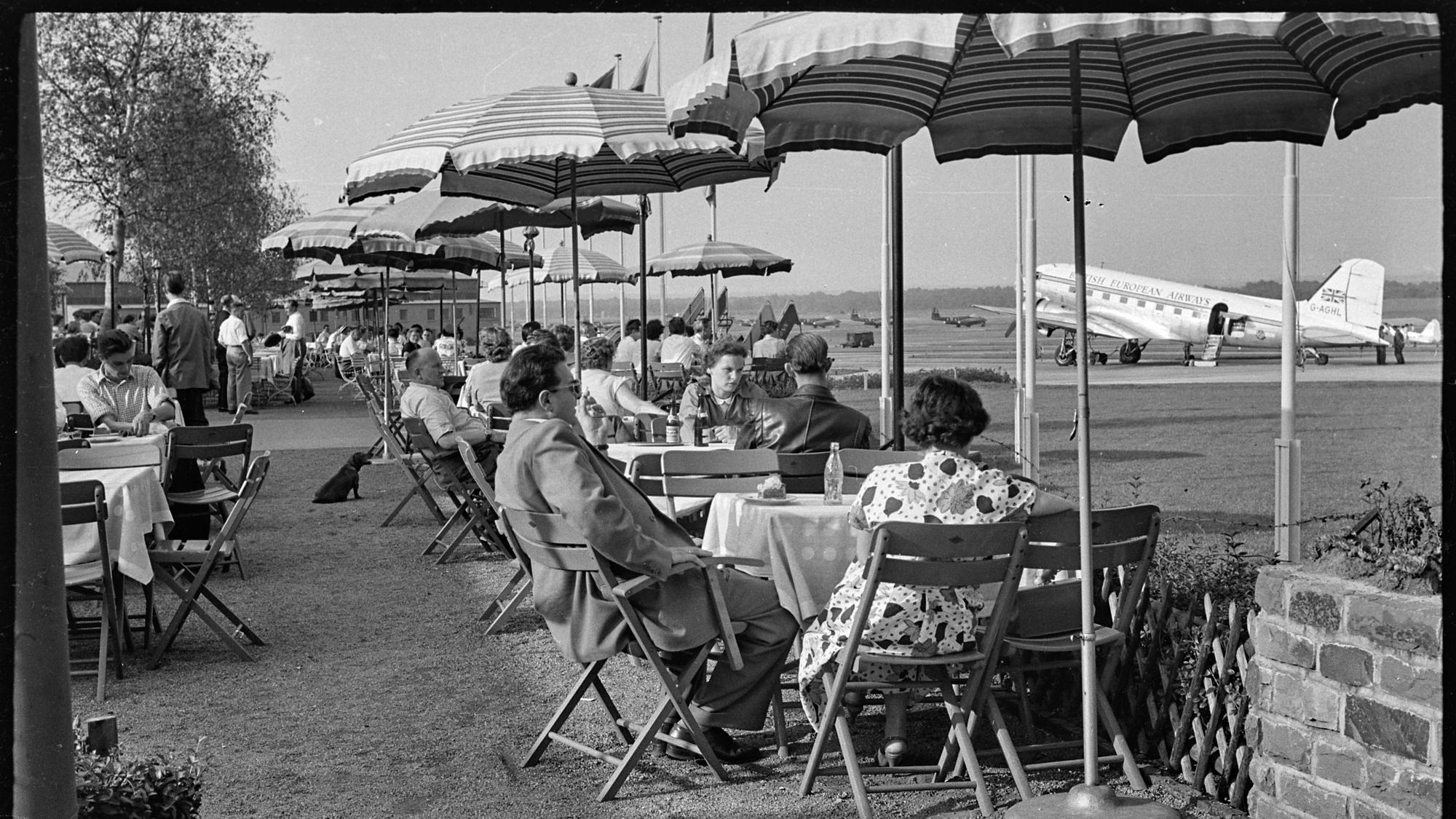 Köln - bei strahlendem Sommerwetter genießen Sonntags-Ausflügler unter Sonnenschirmen auf der Terrasse des Köln-Bonner Flughafens die Aussicht auf die Start- und Landebahn - Foto vom 21.08.1955