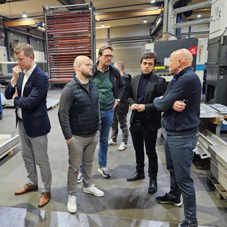 Sechs Männer stehen in einer Werkhalle und sprechen miteinander.