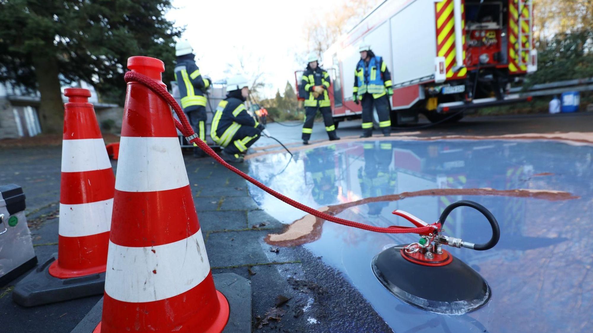 Ein kleiner „See“ mit Löschwasser am Straßenrand, im Hintergrund vier Feuerwehrleute.