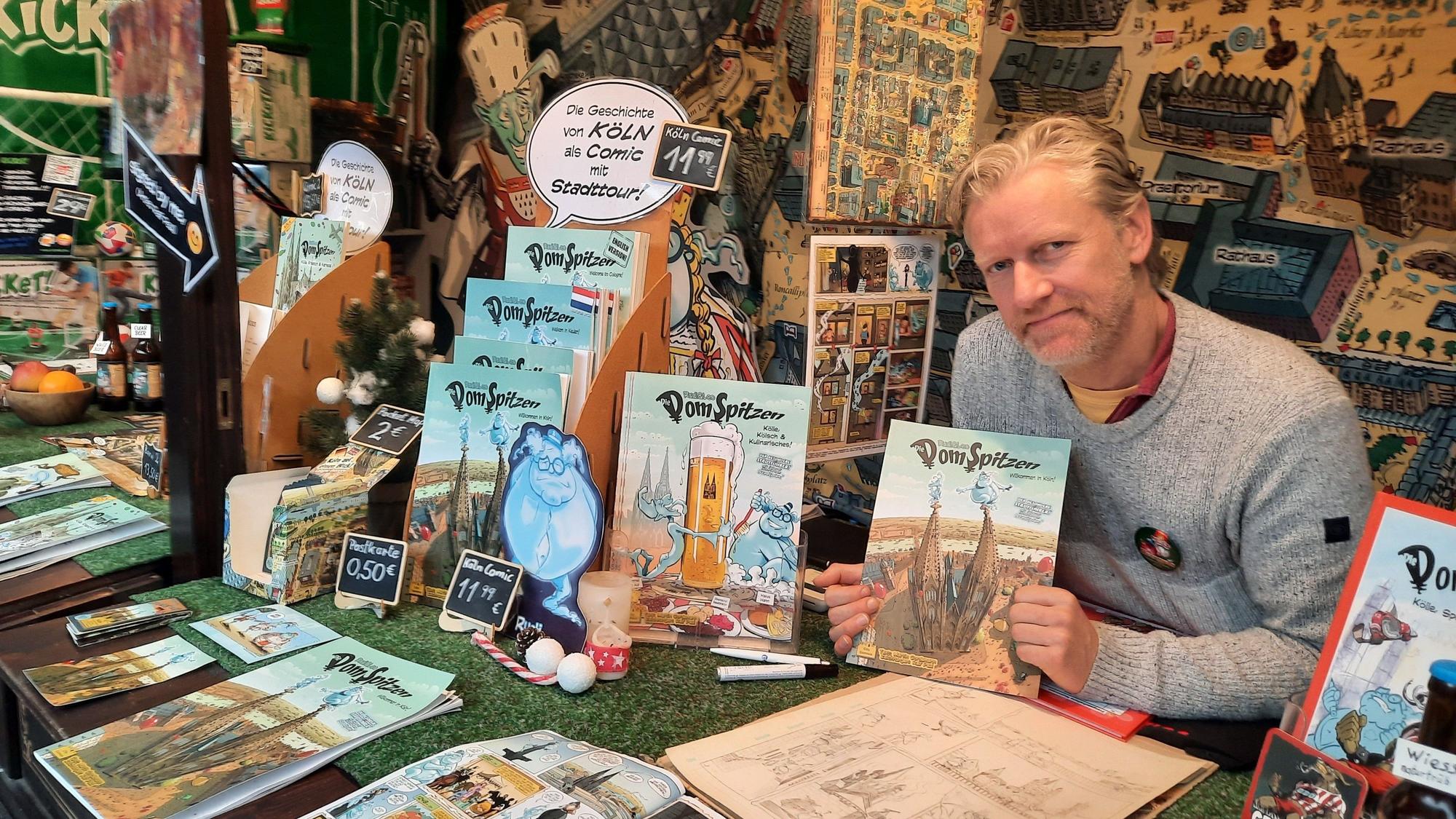 Ralf Paul in seinem Stand auf dem Weihnachtsmarkt. Er hält den Comic "Die Domspitzen" in den Händen.