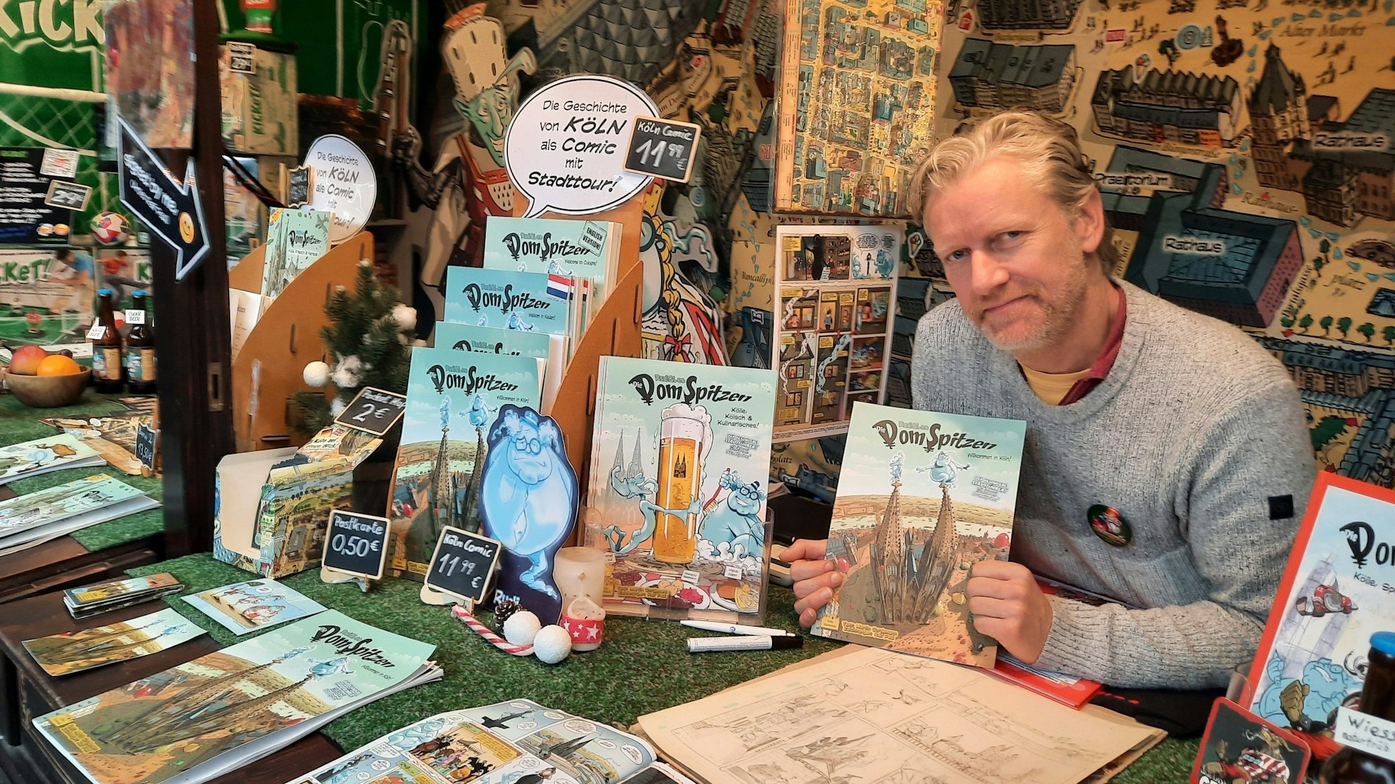 Ralf Paul in seinem Stand auf dem Weihnachtsmarkt. Er hält den Comic "Die Domspitzen" in den Händen.