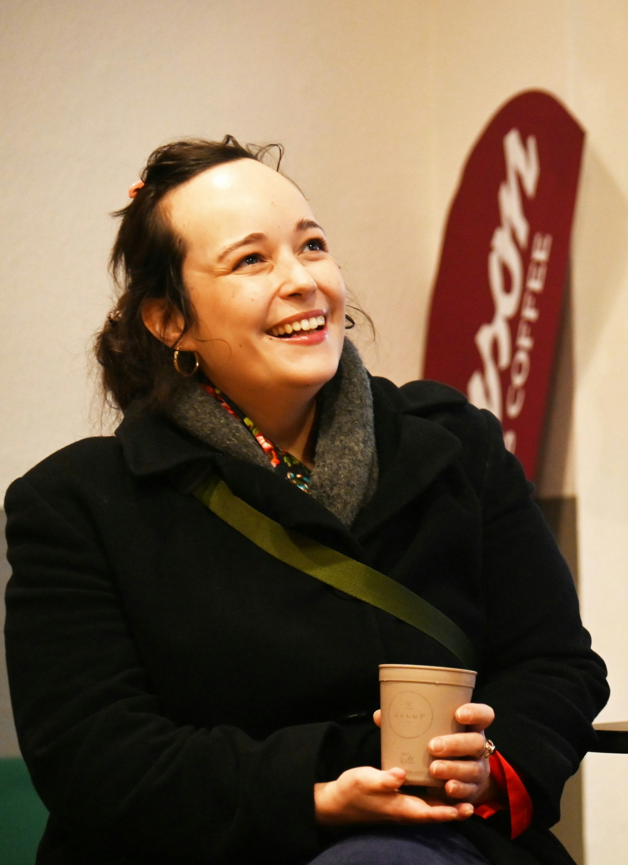 Eine junge Frau hält lachend einen Becher Kaffee in der Hand.