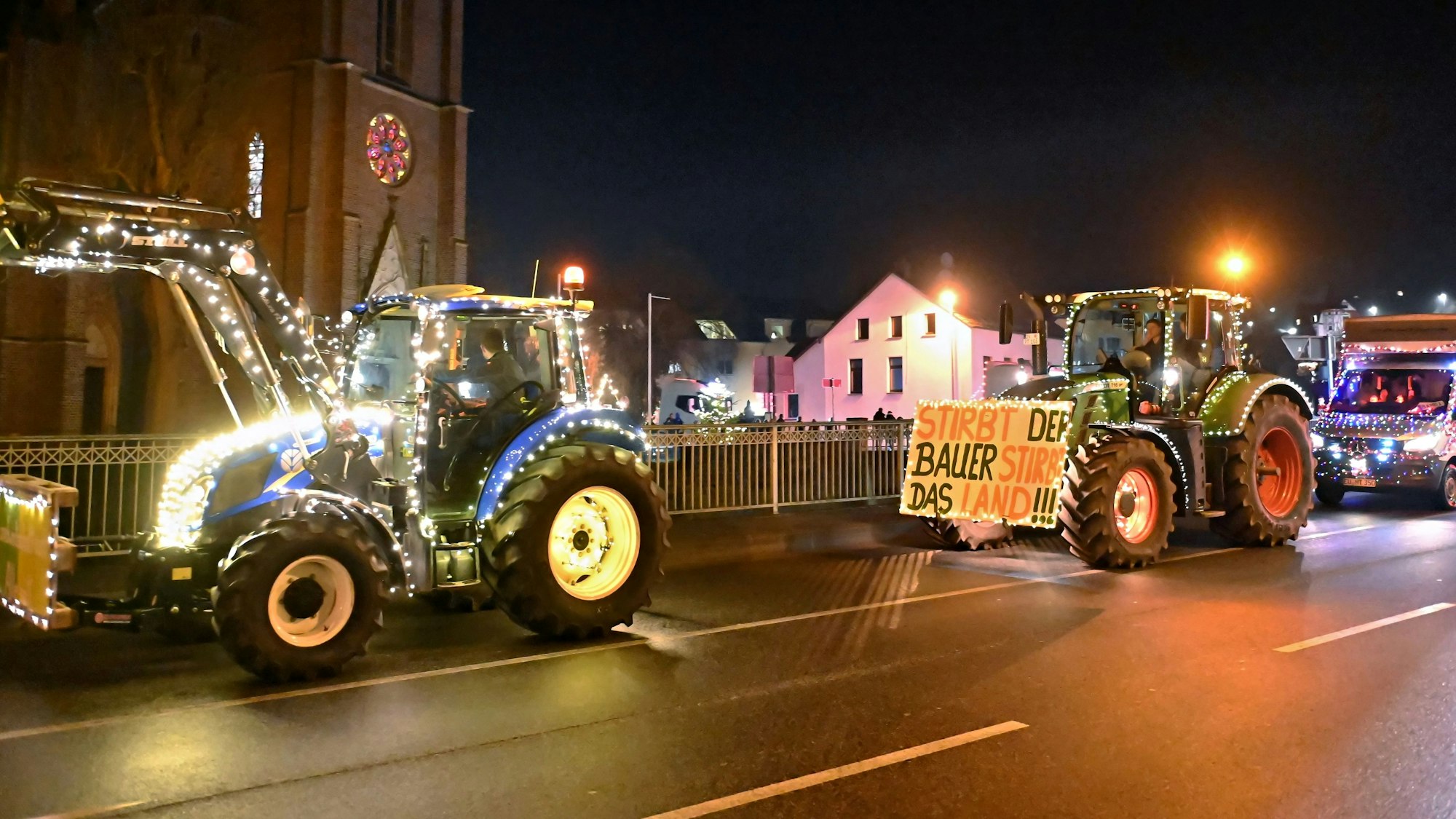 Zwei Traktoren und ein Lkw, die mit Lichterketten geschmückt sind, fahren in der Dunkelheit an der Kirche in Gemünd vorbei.