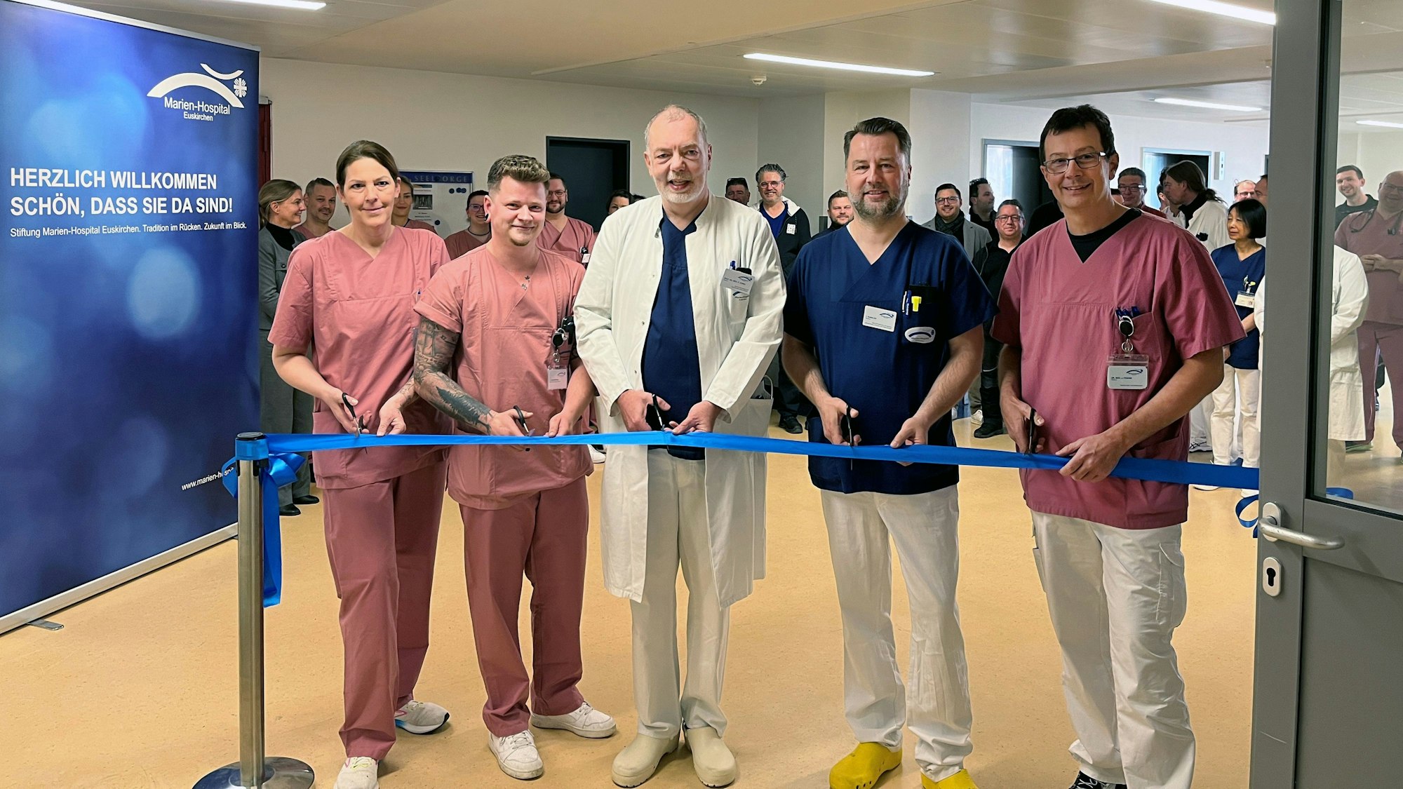 Eine Frau und vier Männer durchschneiden ein blaues Band, hinter dem sie in einer Reihe stehen.