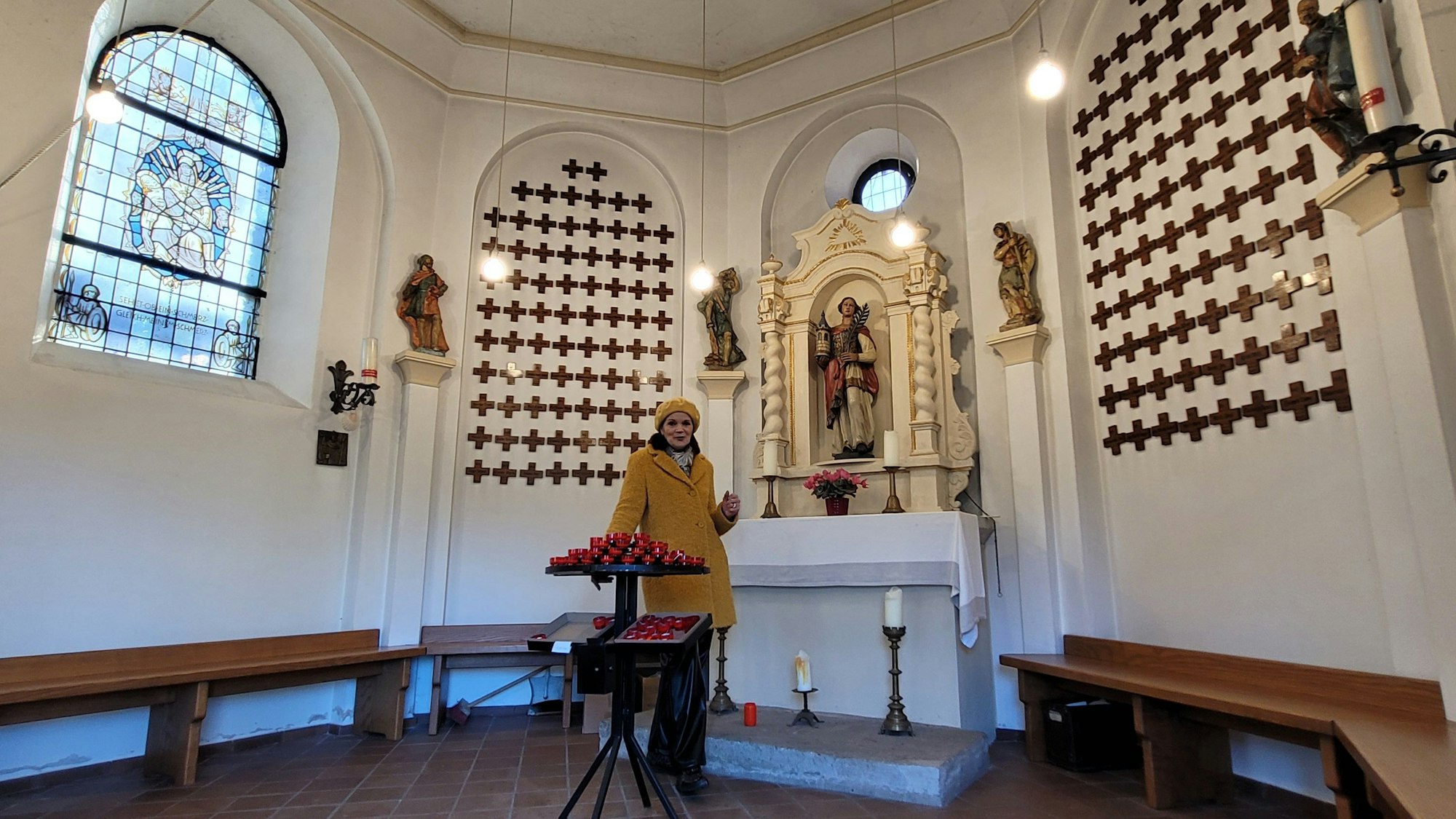 Eine Frau steht in einem achtseitigen Raum, hinter ihr steht ein barocker Altar mit einer Figur der Heiligen Barbara, an den Wänden hängen Holzkreuze.
