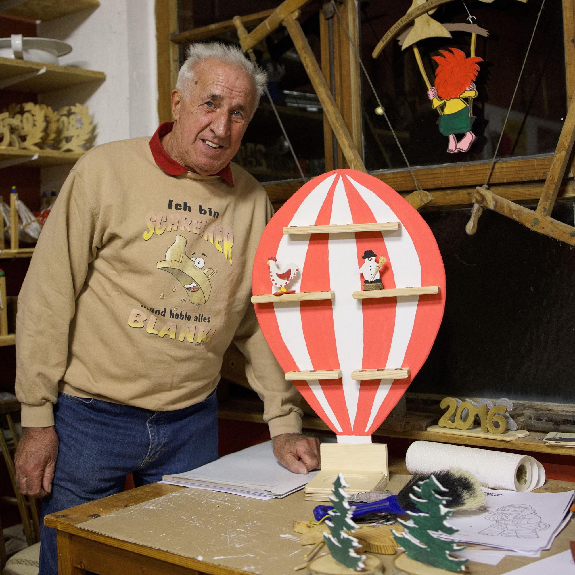 Der Schreiner steht in seiner Werkstatt. Auf dem Tisch steht ein aus Holz geschnittener Heißluftballon, im Hintergrund schaukelt eine Pumuckl-Figur.